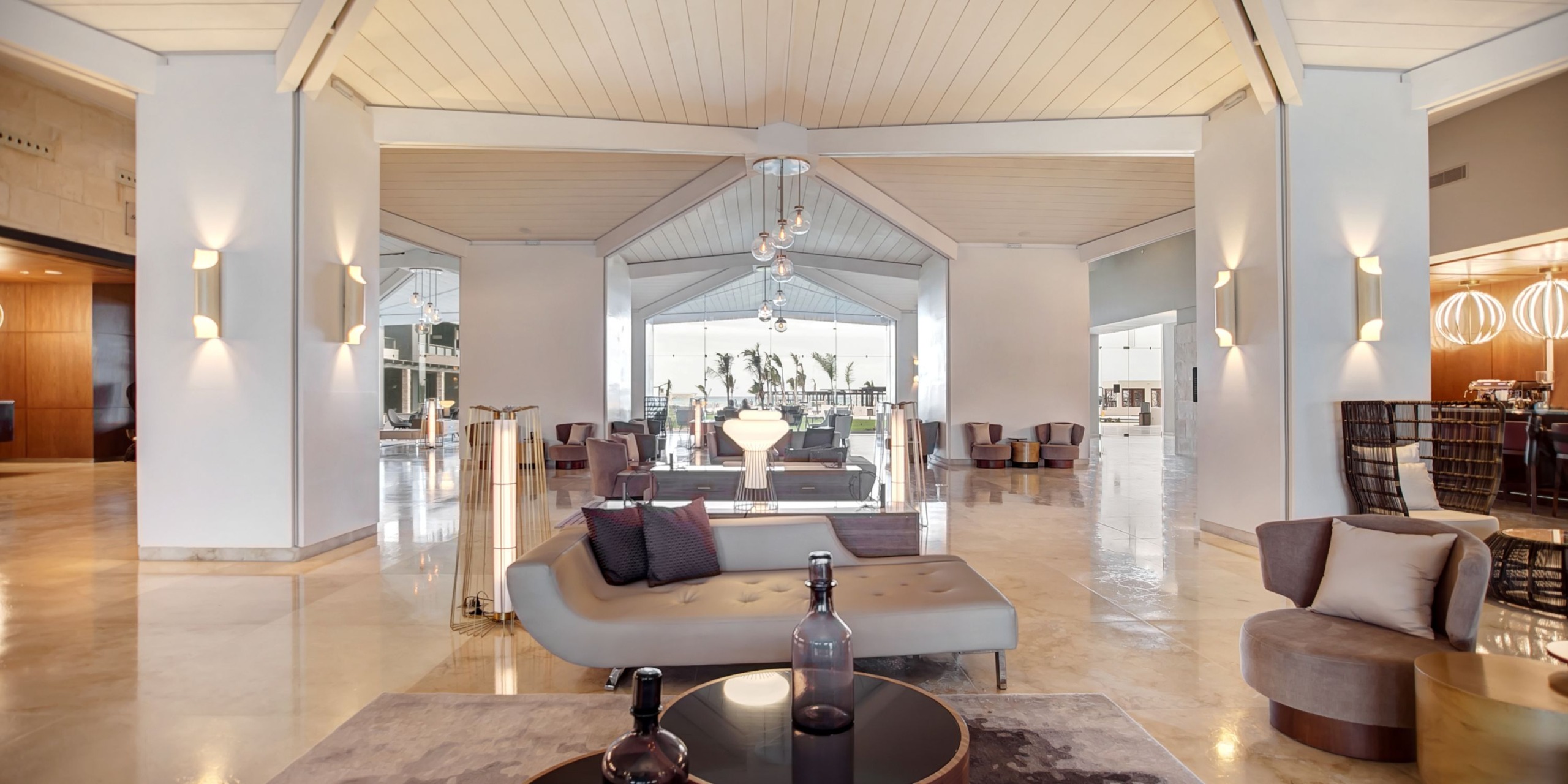 The lobby at Royalton Blue Waters is spacious, featuring modern furniture like a gray sofa and round tables. Large windows provide a view of the outdoor area, where palm trees can be seen in the distance.