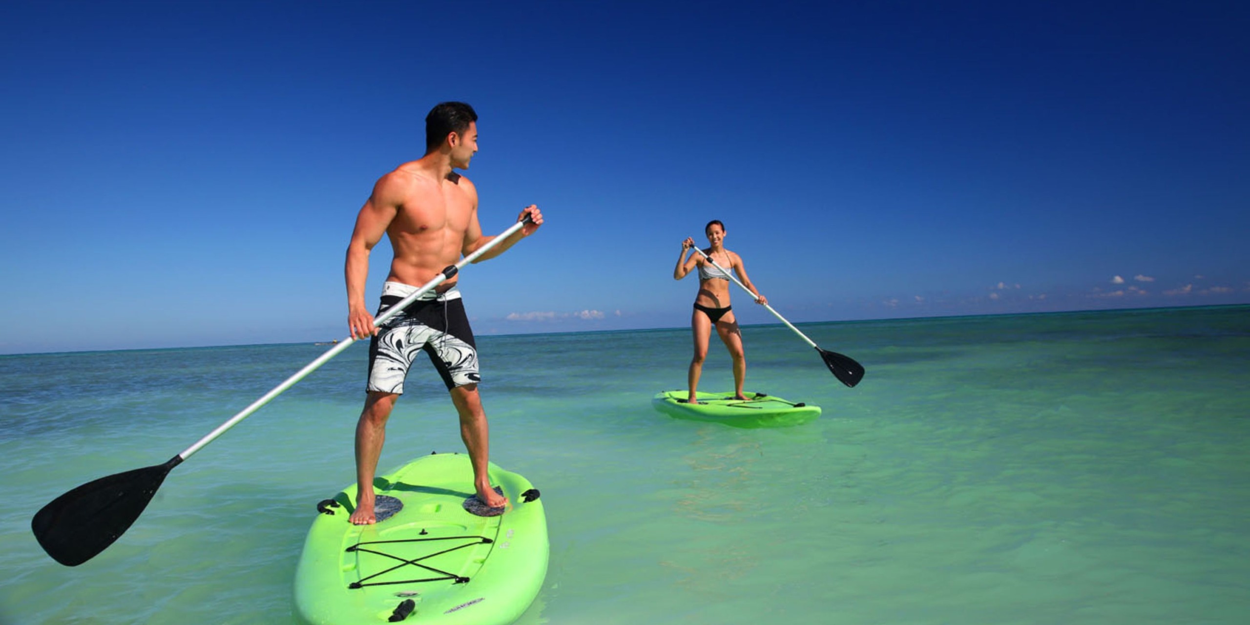 Two people are paddleboarding on Royalton Blue Waters in clear, turquoise water under a blue sky.