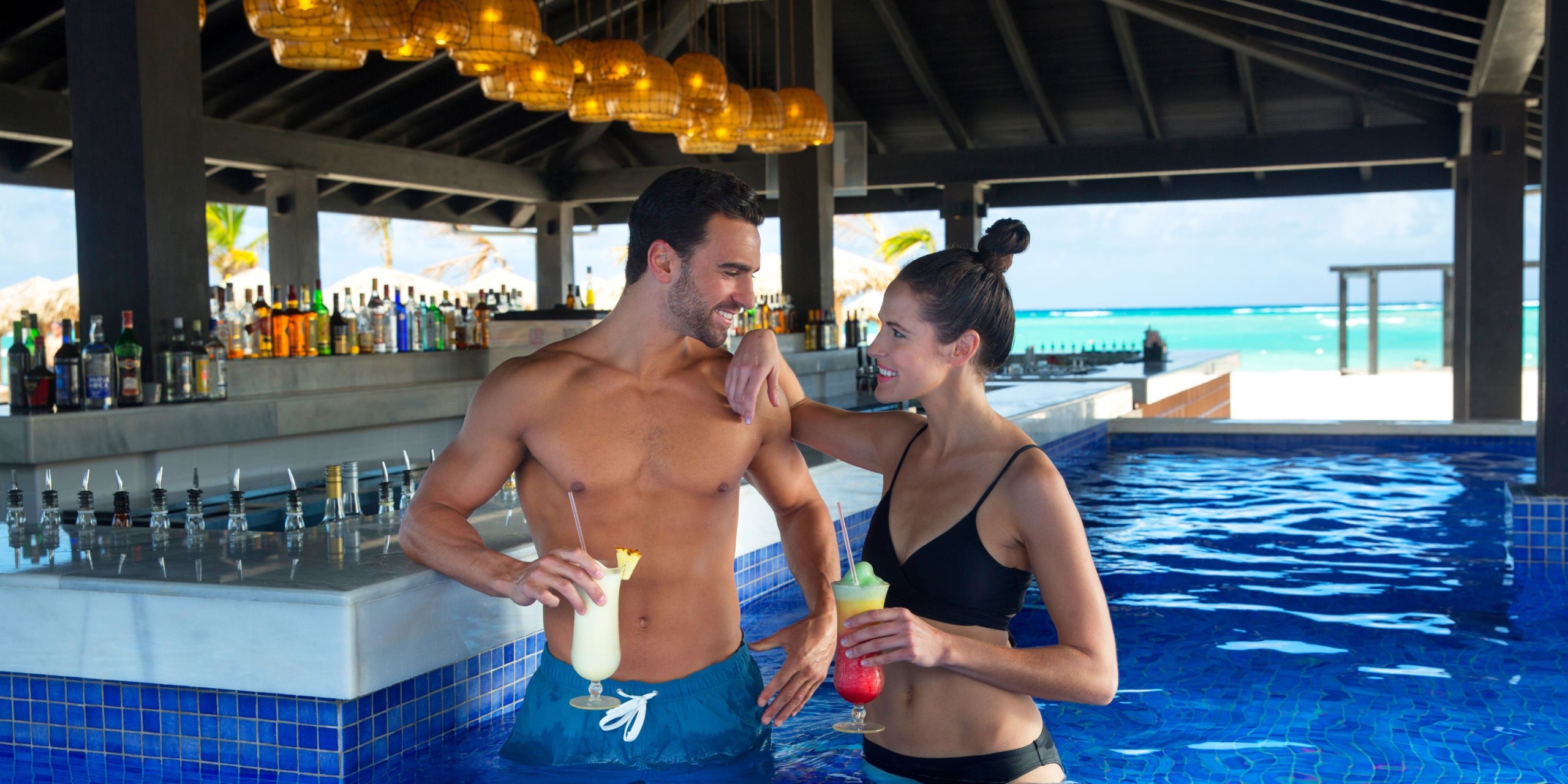 At the Royalton Blue Waters, a man and woman in swimwear enjoy themselves at the poolside bar, each holding a tropical drink and sharing smiles.
