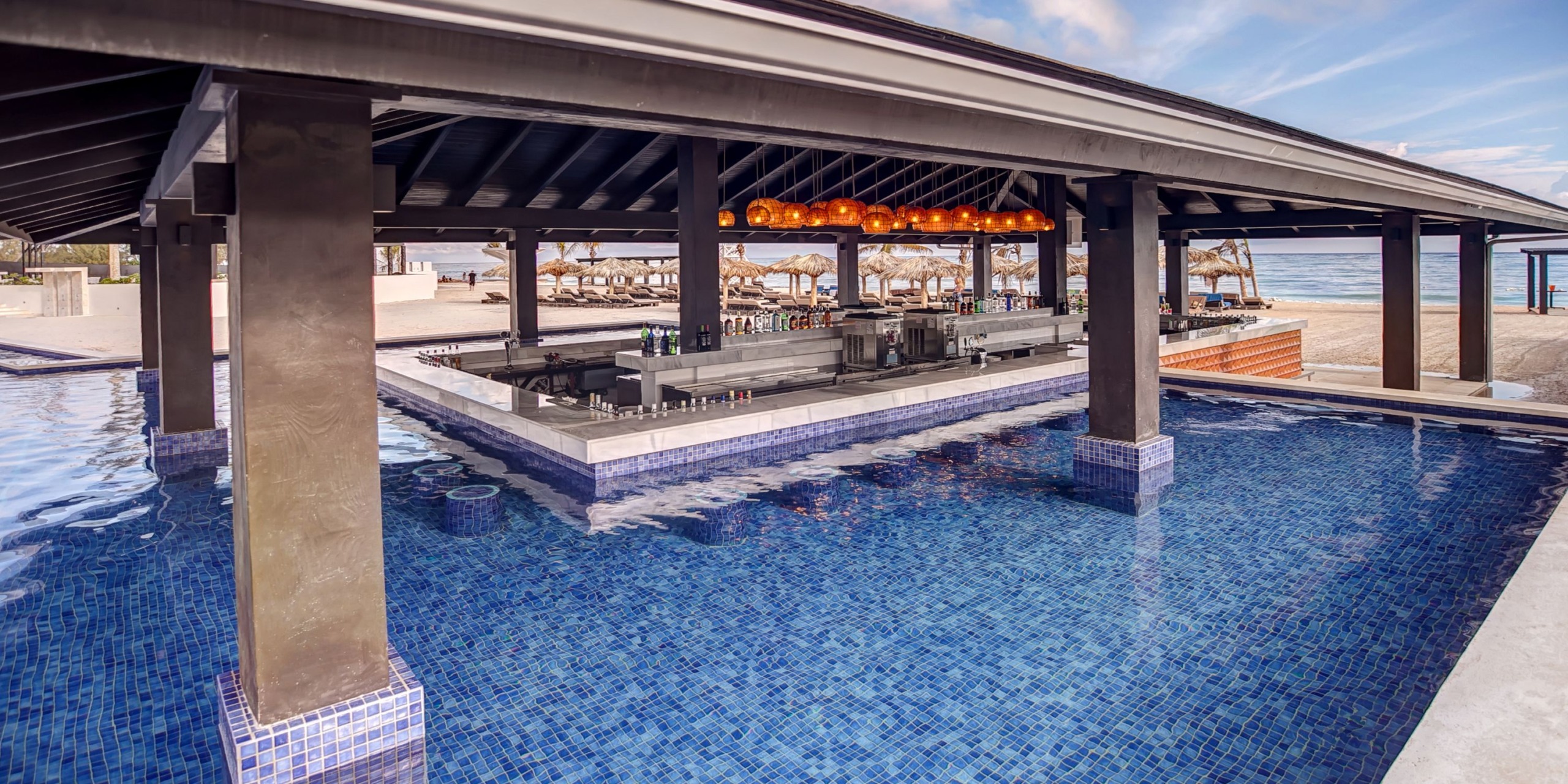 The Royalton Blue Waters features a poolside bar beneath a thatched roof, encircled by water with available seating and tables, and offering views of the beach and ocean in the background.