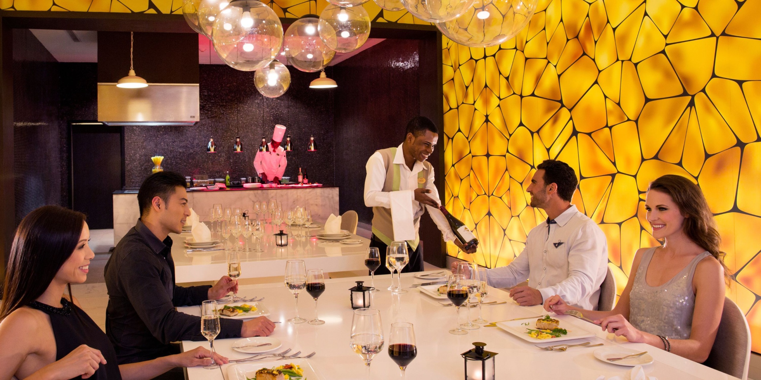 At Royalton Blue Waters, a waiter pours wine for guests seated at a modern table in front of a yellow geometric wall. Four people are enjoying a meal with elegantly set wine glasses.