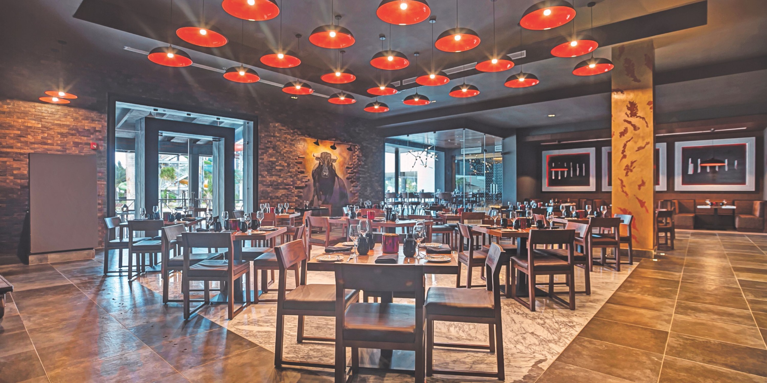 An upscale dining space featuring wooden tables and chairs, complemented by red circular ceiling lights and large windows in the backdrop, reminiscent of the Royalton Blue Waters ambiance.