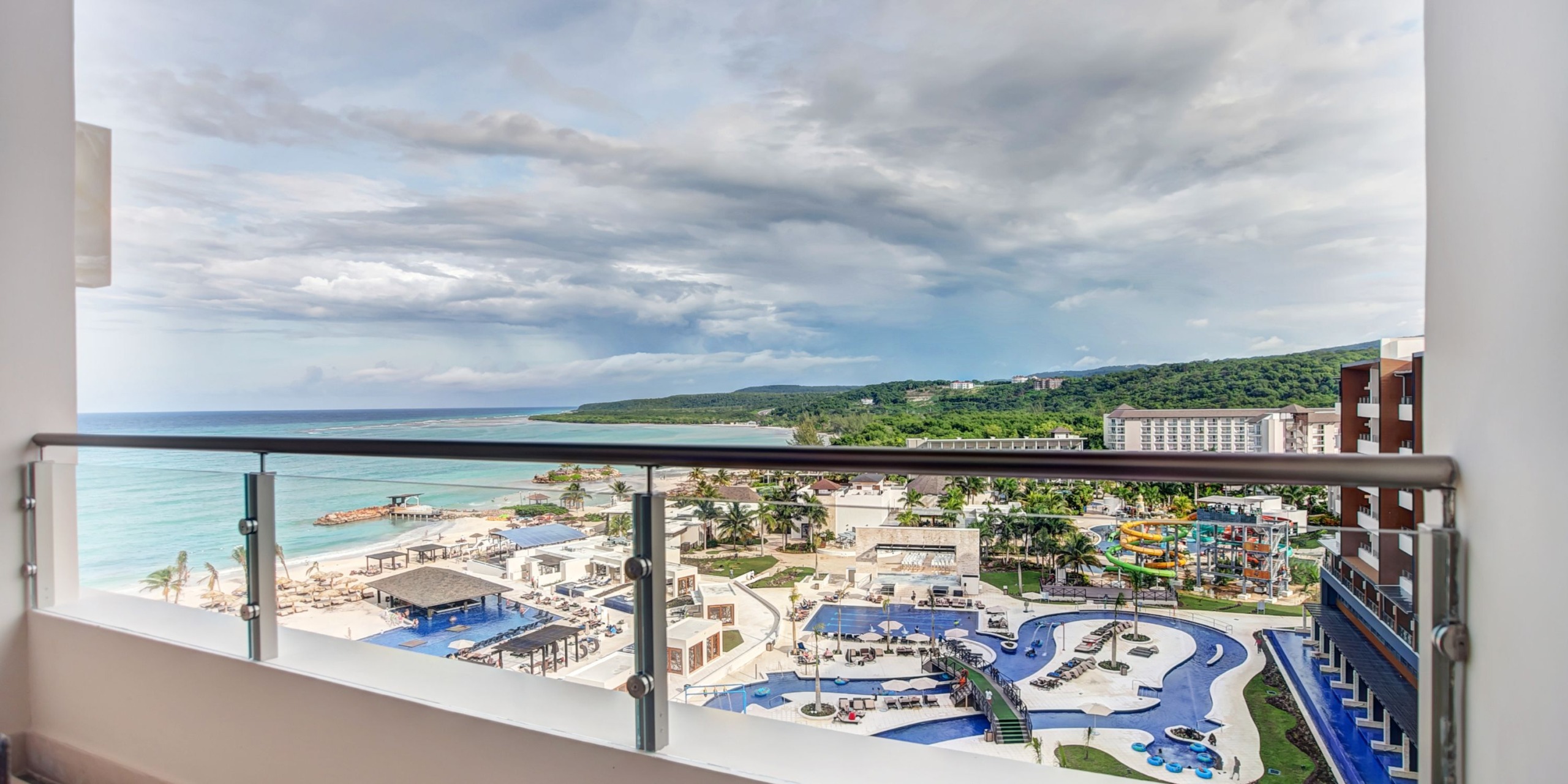 Gaze from a balcony overlooking the Royalton Blue Waters resort, featuring a pool, water park, and beach, all set against a stunning ocean backdrop with a cloudy sky.