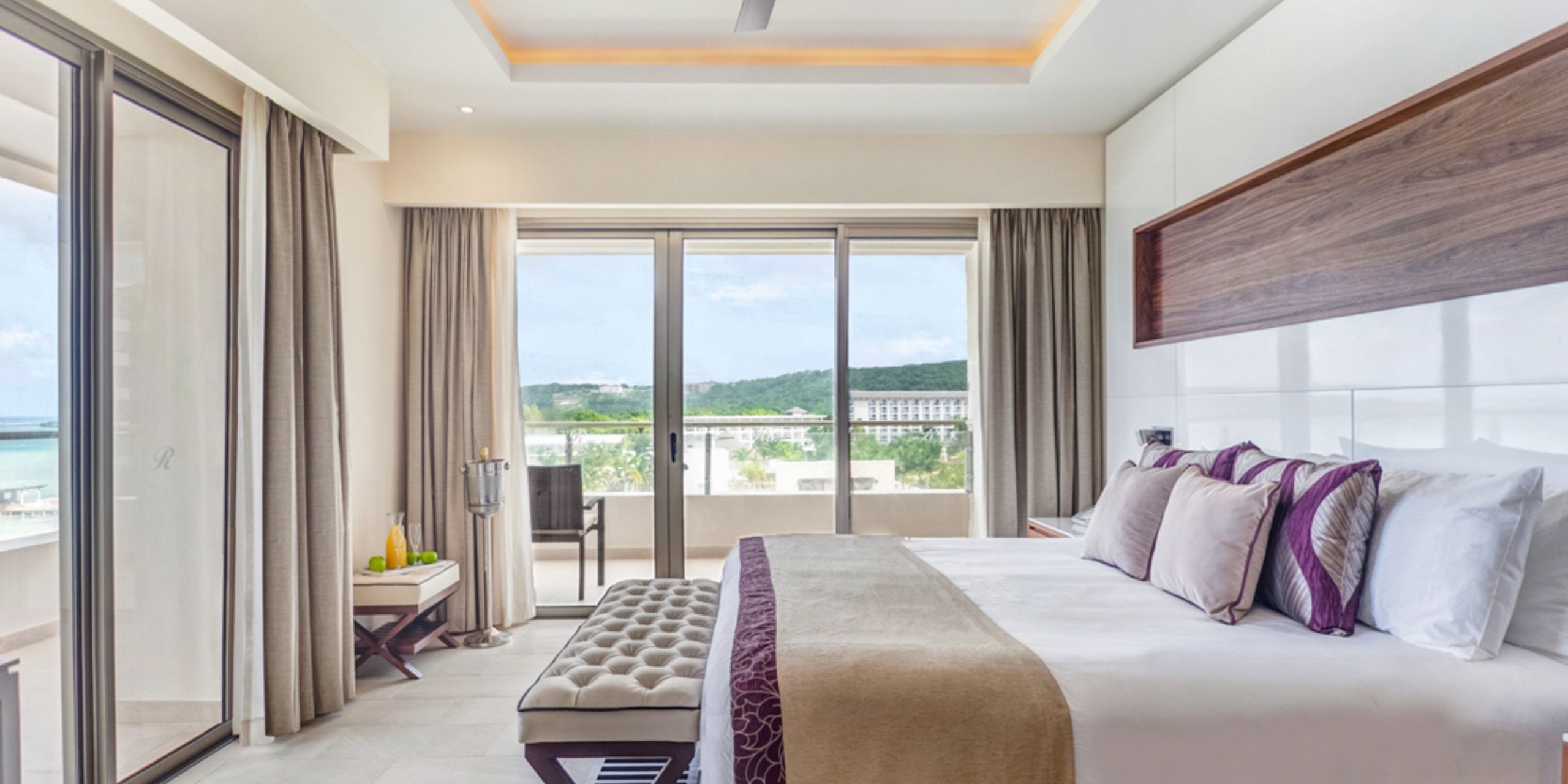 Enjoy a stay in a modern hotel room at Royalton Blue Waters, featuring a large bed adorned with purple accents, floor-to-ceiling windows that provide stunning scenic views, and a cozy corner with a small table complete with a bottle and glasses.