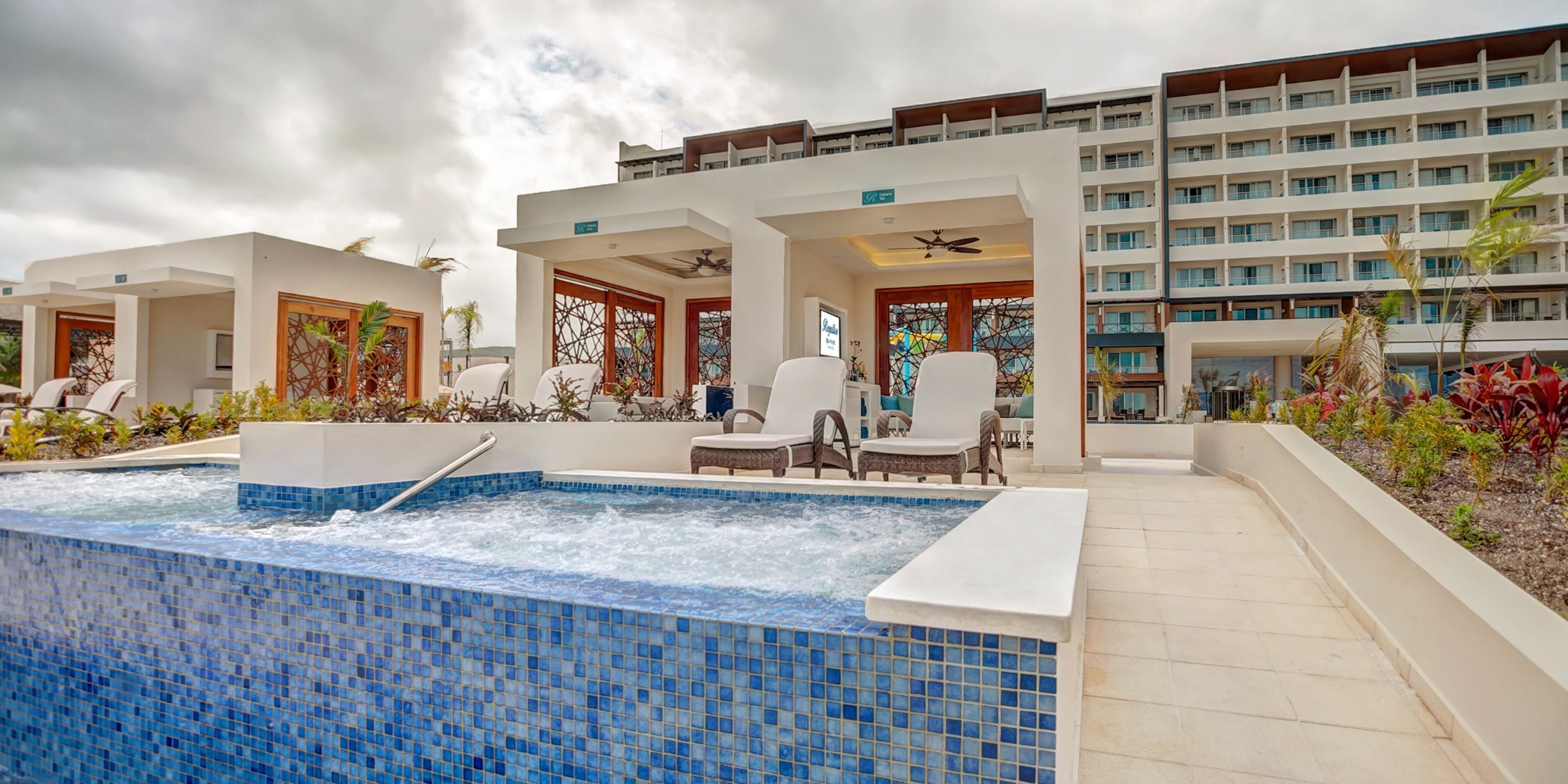 The Royalton Blue Waters is a contemporary hotel that boasts a tiered pool area complete with lounge chairs and cabanas, all situated against the backdrop of a multi-story building enveloped by a cloudy sky.