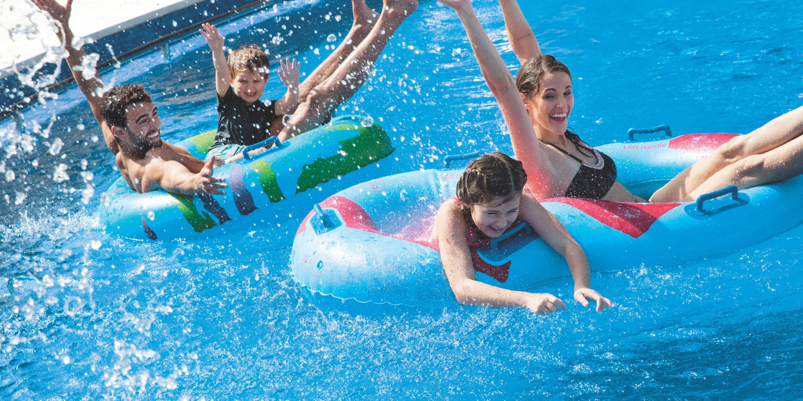 A group of four people glides down a water slide on inflatable tubes, making a splash as they plunge into the pool at Royalton Blue Waters.