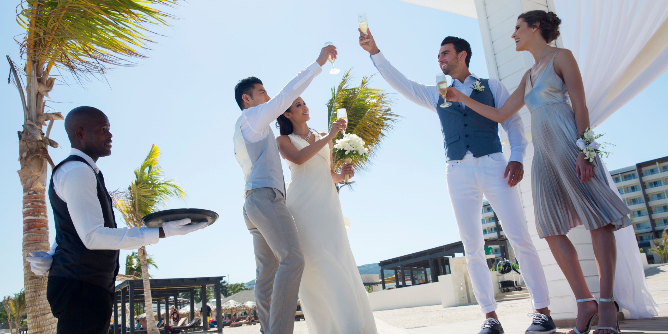 A wedding group at Royalton Blue Waters toasts with champagne outdoors near palm trees, while a server holds a tray nearby.