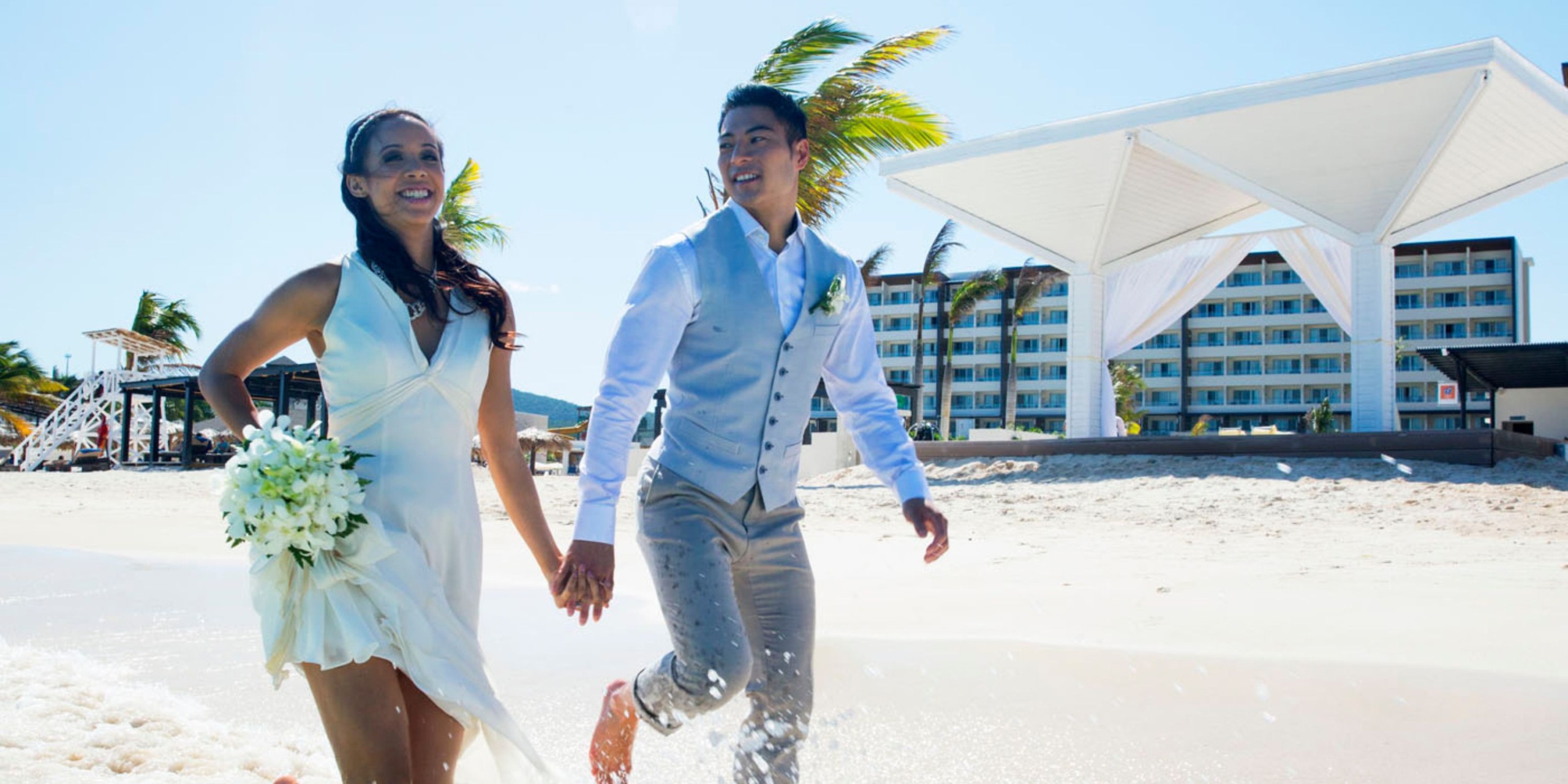 A smiling couple in wedding attire holds hands, walking along a sunny beach with the Royalton Blue Waters hotel in the background.