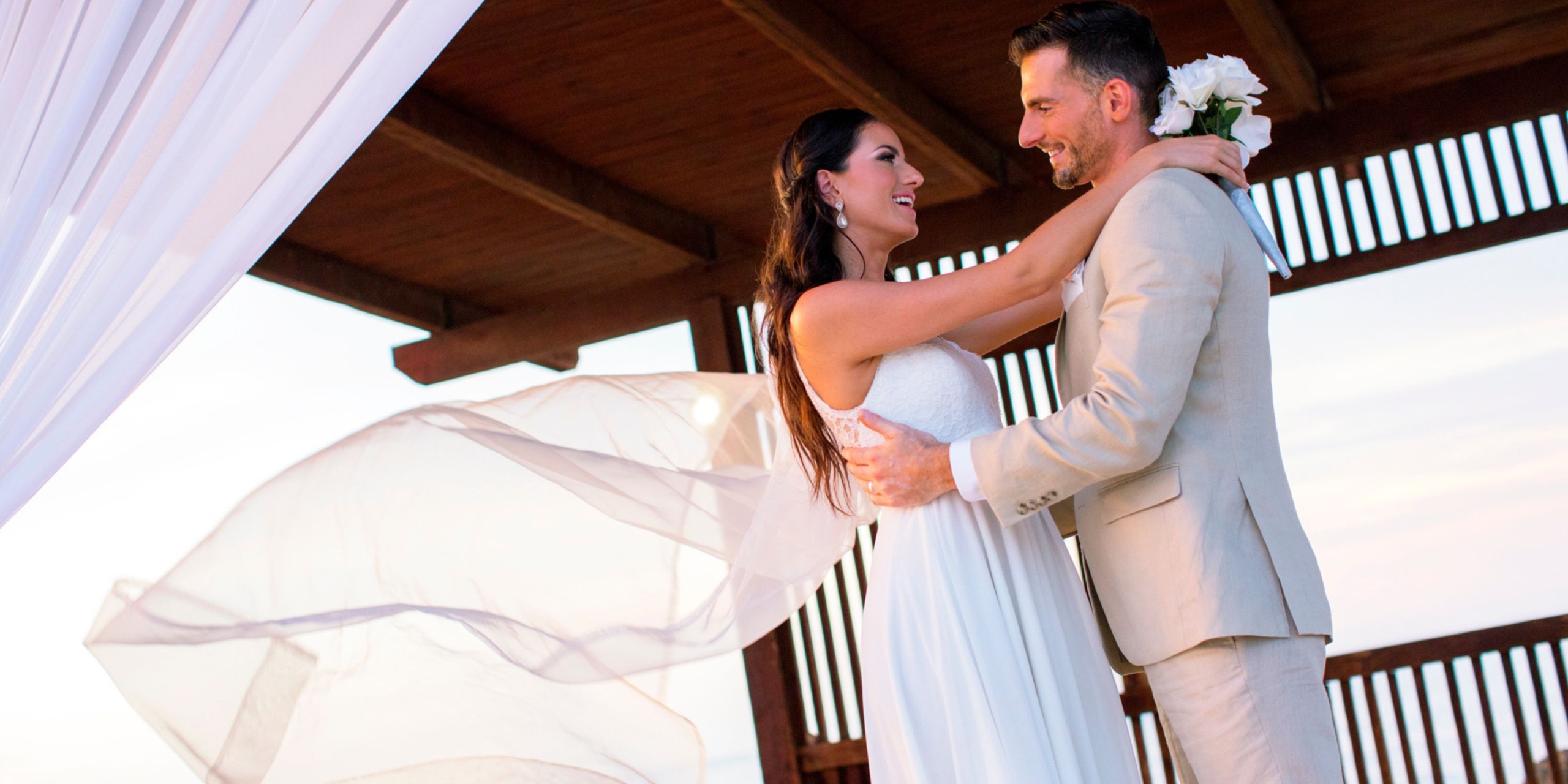 The bride and groom embrace under a wooden canopy at Royalton Blue Waters, with the bride's veil flowing in the breeze.