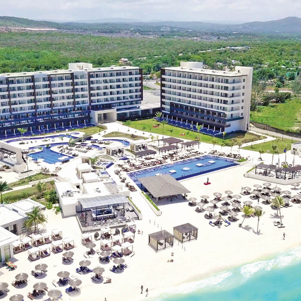 Aerial view of Royalton Blue Waters, a modern beachfront resort, perfect for destination weddings, with multiple swimming pools, loungers, and a lush green landscape in the background.