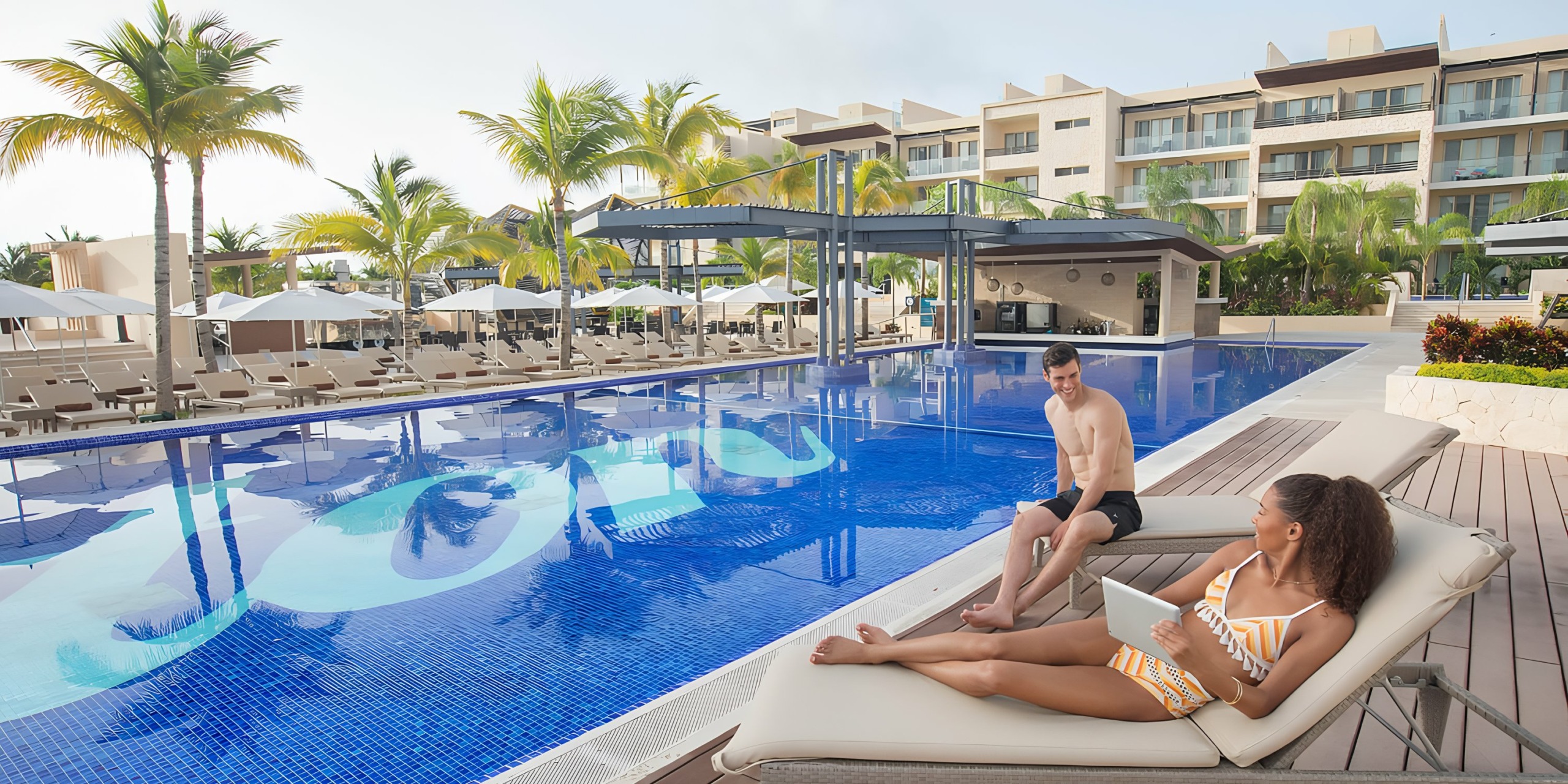 A man sitting on the edge of an outdoor pool at the Royalton Riviera Cancun, looking at a woman lying on a sun lounger at the Royalton Riviera Cancun luxury resort.