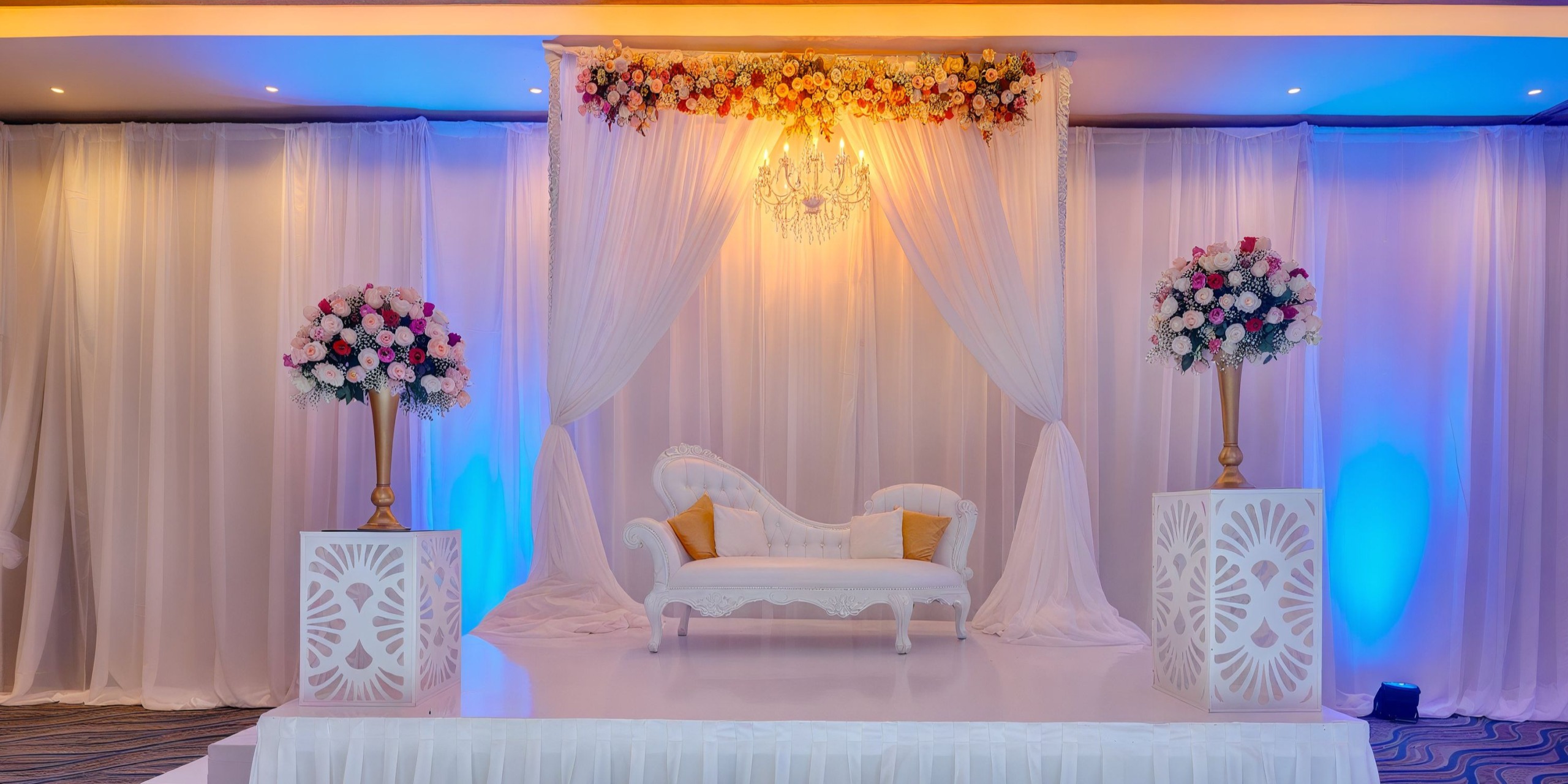 A white lounge sofa graces a decorated stage adorned with sheer curtains, floral arrangements, and an overhead chandelier, capturing the elegance of a Royalton Riviera Cancun setting.