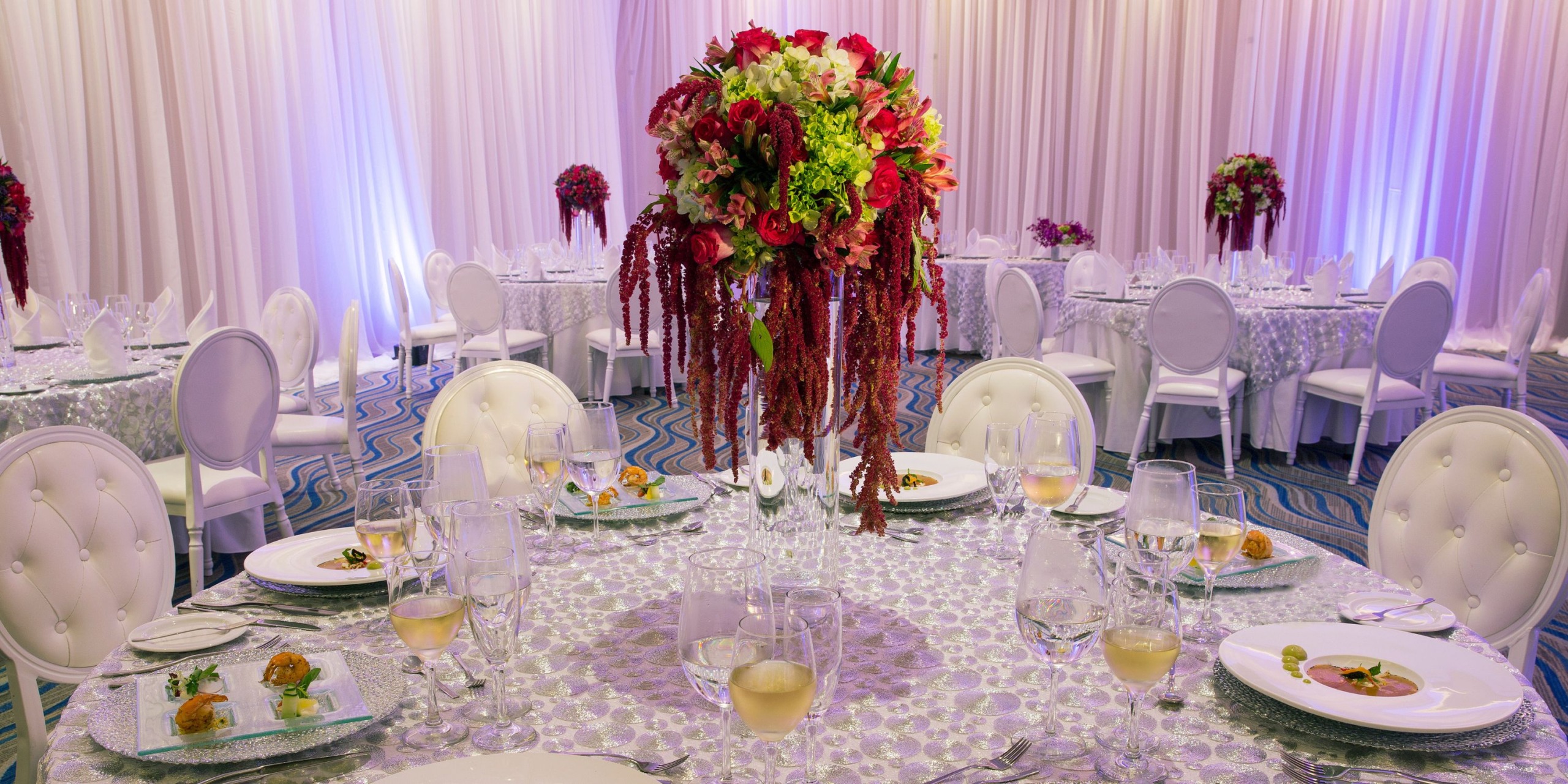 Celebrate your wedding at the stunning Royalton Riviera Cancun, where our refined banquet hall offers round tables with beautiful floral centerpieces, elegant white chairs, and precisely arranged silverware for a memorable dining experience.