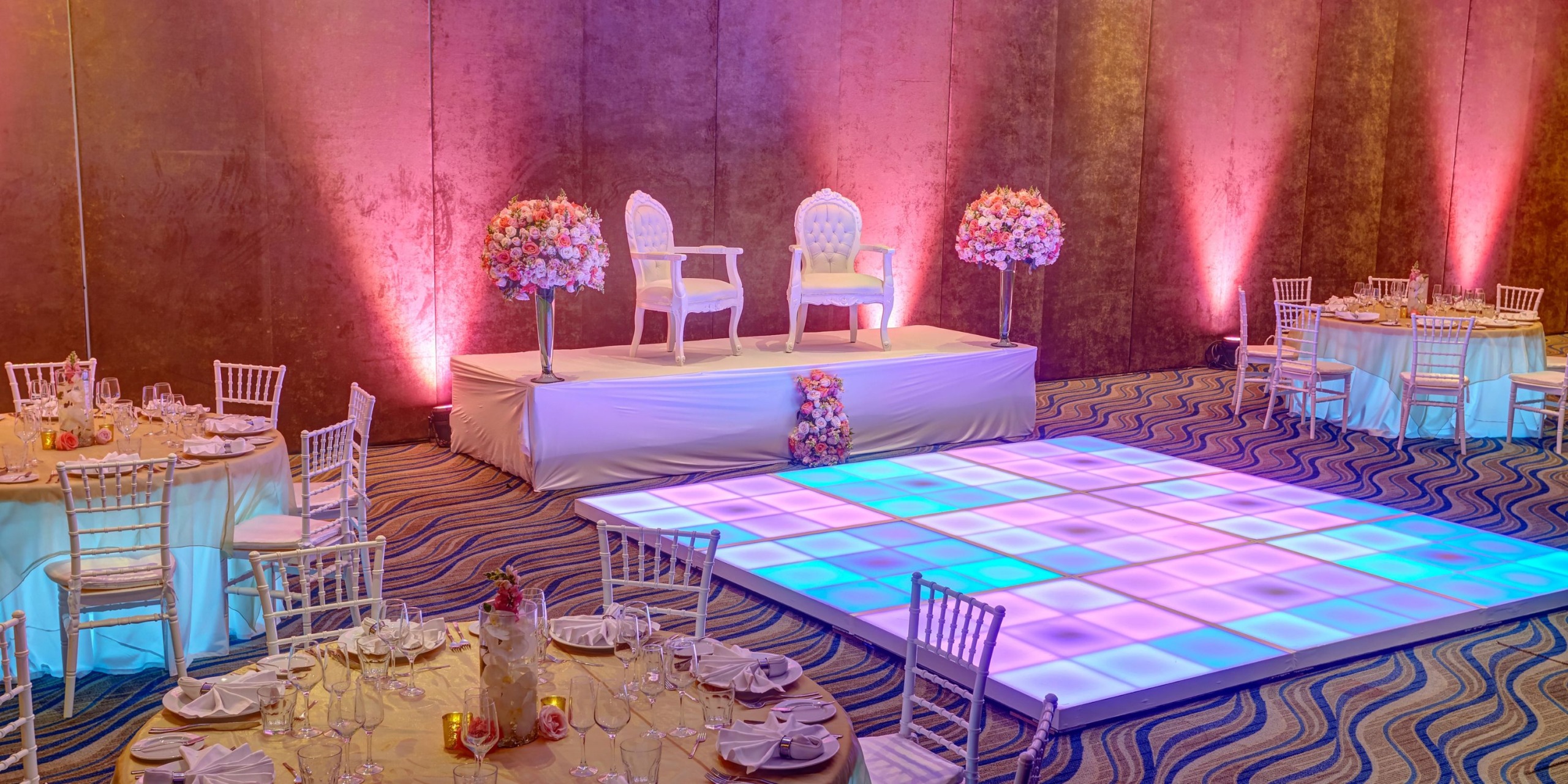 The Royalton Riviera Cancun provides a beautifully decorated wedding venue that includes an elegant stage with two sophisticated chairs, exquisite floral arrangements, round tables set with fine tableware, and a brilliantly illuminated dance floor.