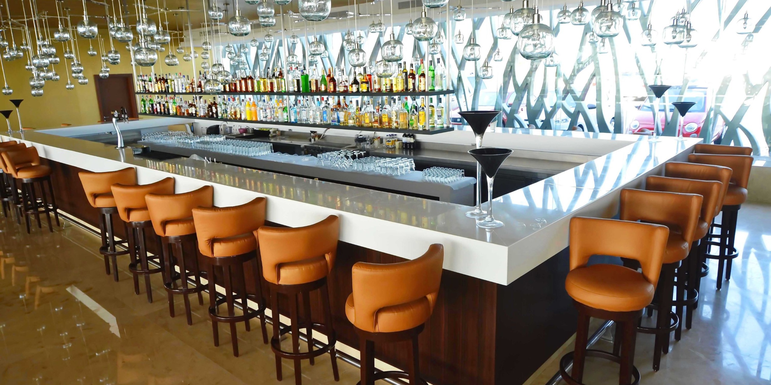 The Royalton Riviera Cancun features a chic bar with a U-shaped counter, complemented by leather stools and pendant lights, creating an ideal atmosphere to relax after your wedding. The back wall showcases shelves filled with bottles, encouraging guests to celebrate in style.