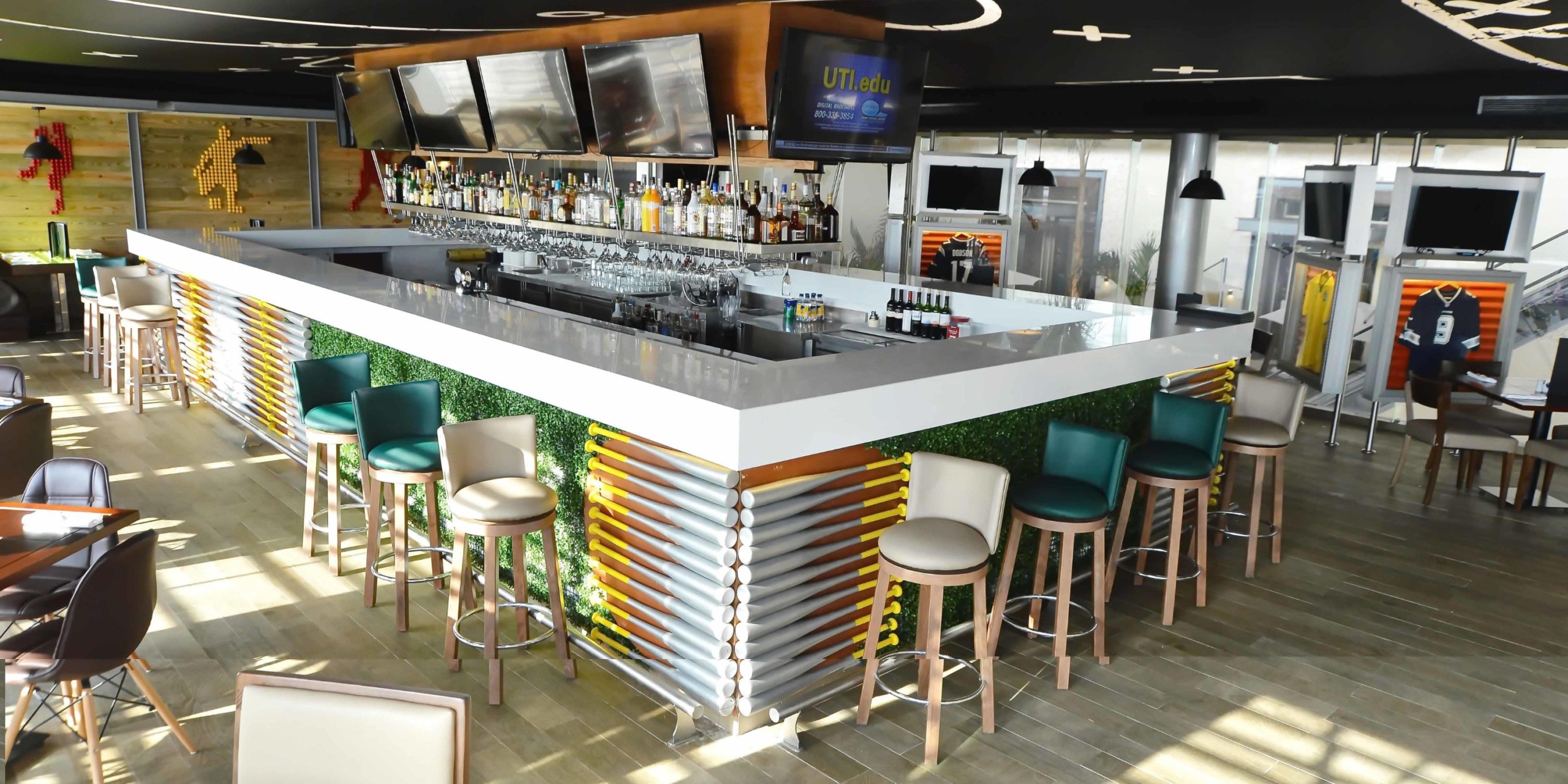 A stylish bar, inspired by the elegance of the Royalton Riviera Cancun, features a large square counter adorned with sleek metal details and lush greenery accents. Comfortable bar stools beckon guests to unwind, while multiple TV screens guarantee entertainment in this contemporary ambiance.