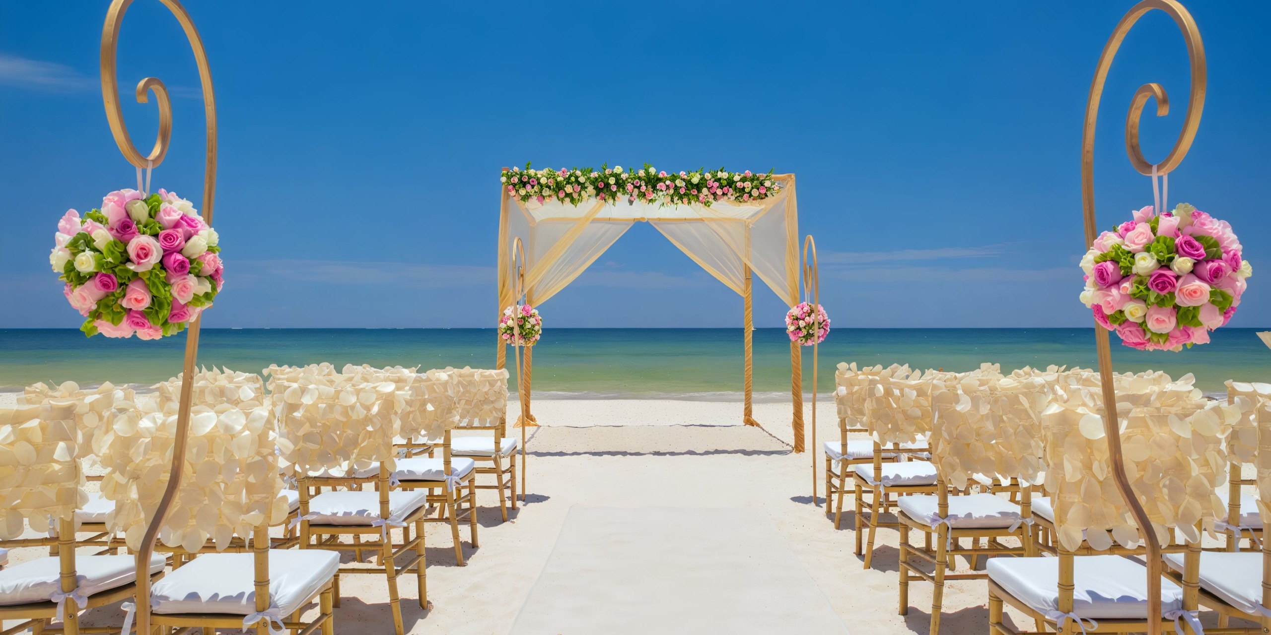 The wedding setup at the Royalton Riviera Cancun showcases a breathtaking floral arch, complemented by white chairs and delicate pink flower arrangements, beautifully arranged on the beach with the ocean as a backdrop under a clear blue sky.