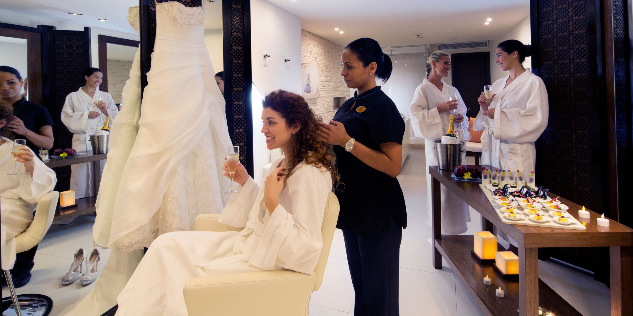 Seated in a salon chair, a woman enjoys her drink while having her hair styled for her Royalton Riviera Cancun wedding. Nearby, a wedding dress awaits as other women in robes chat and savor snacks at an elegantly candlelit table.