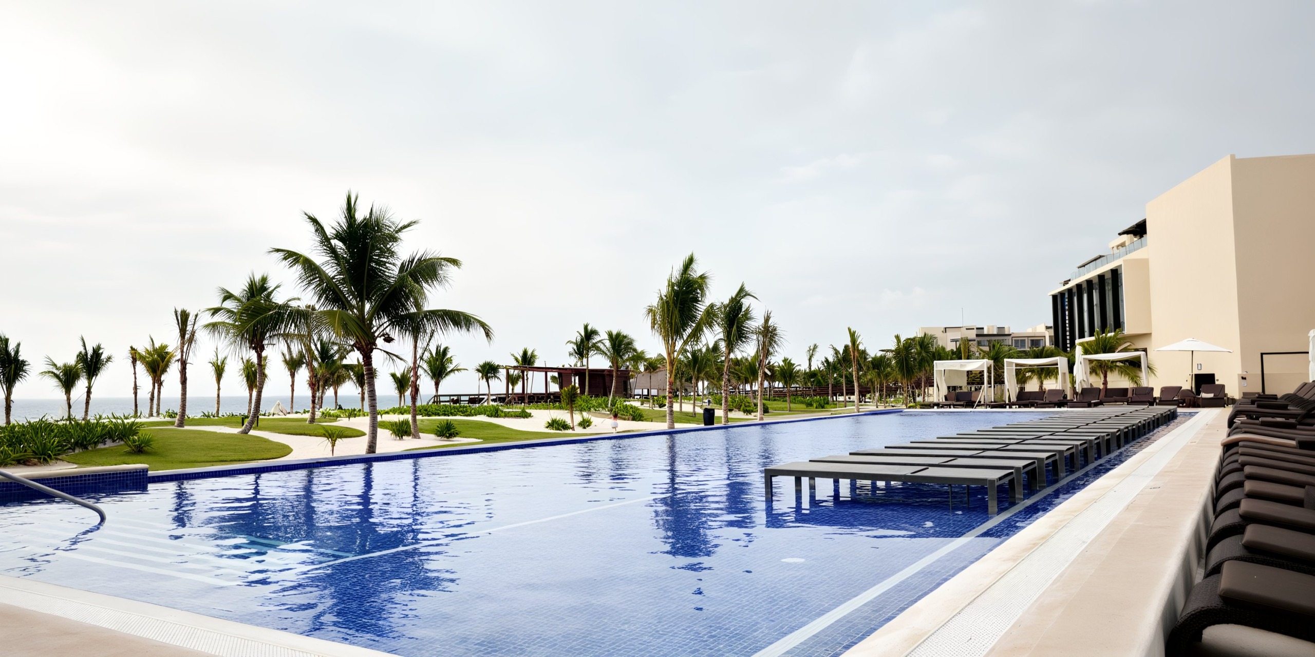 At Royalton Riviera Cancun, the vast outdoor swimming pool creates an unforgettable wedding ambiance, beautifully framed by majestic palm trees and inviting lounge chairs, with a breathtaking building visible in the background.