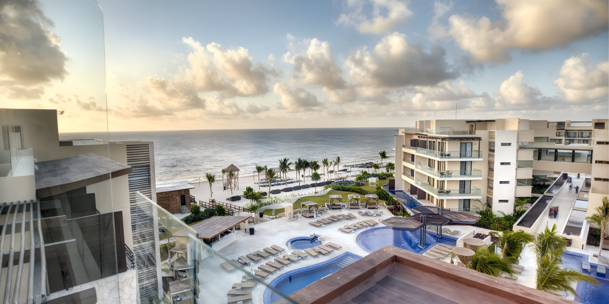 Aerial view of Royalton Riviera Cancun, a coastal resort featuring contemporary architecture, pool areas, and lounge chairs nestled among palm trees, creating a perfect wedding backdrop. The resort boasts stunning views of a sandy beach and the ocean beneath a partly cloudy sky.