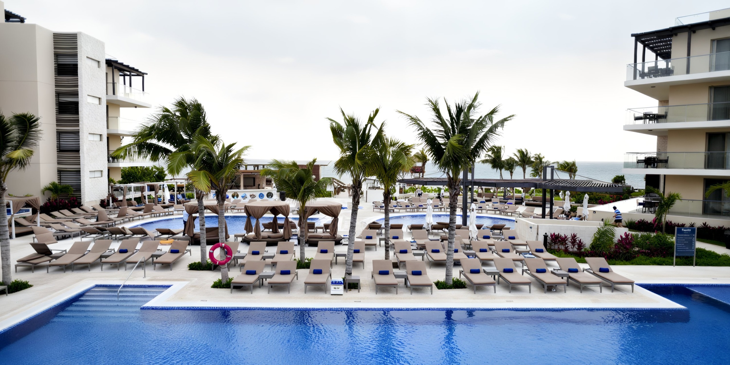 The Royalton Riviera Cancun provides a romantic wedding setting with an outdoor resort pool area, complete with rows of lounge chairs, palm trees, and cabanas. The white buildings surrounding the area frame a stunning ocean view under a cloudy sky.