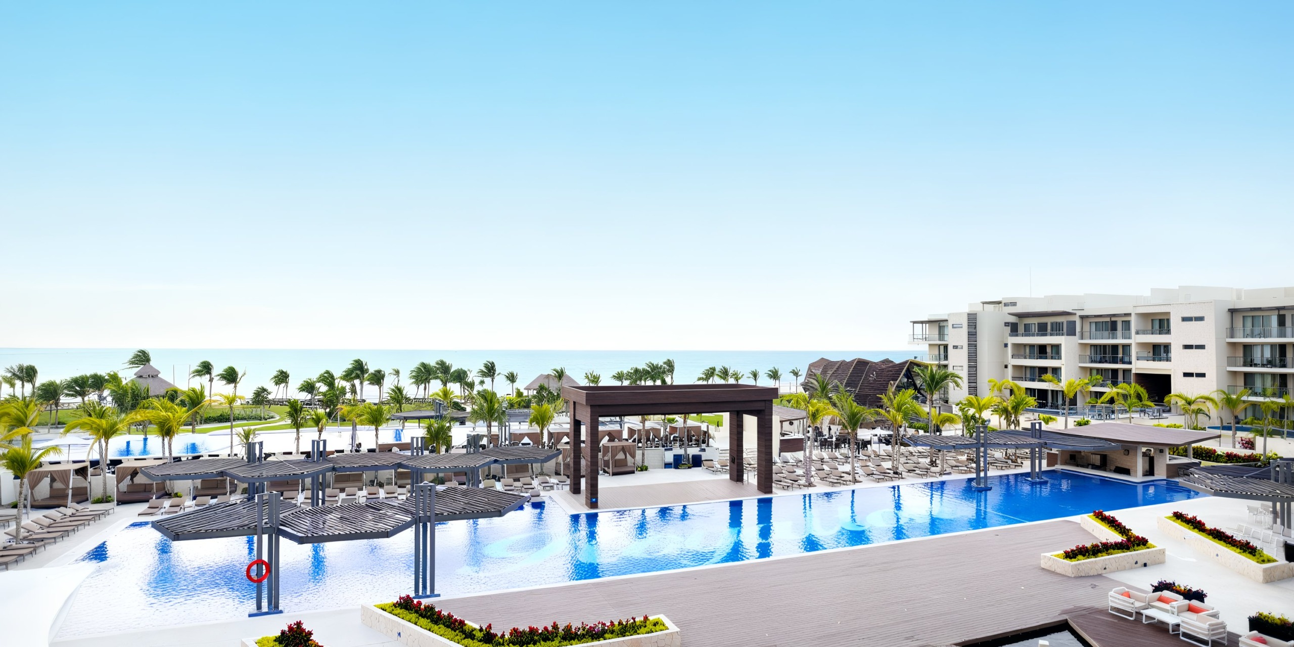 The Royalton Riviera Cancun boasts a spacious resort pool with shaded lounge areas, surrounded by palm trees, offering an ocean view beneath a clear blue sky.