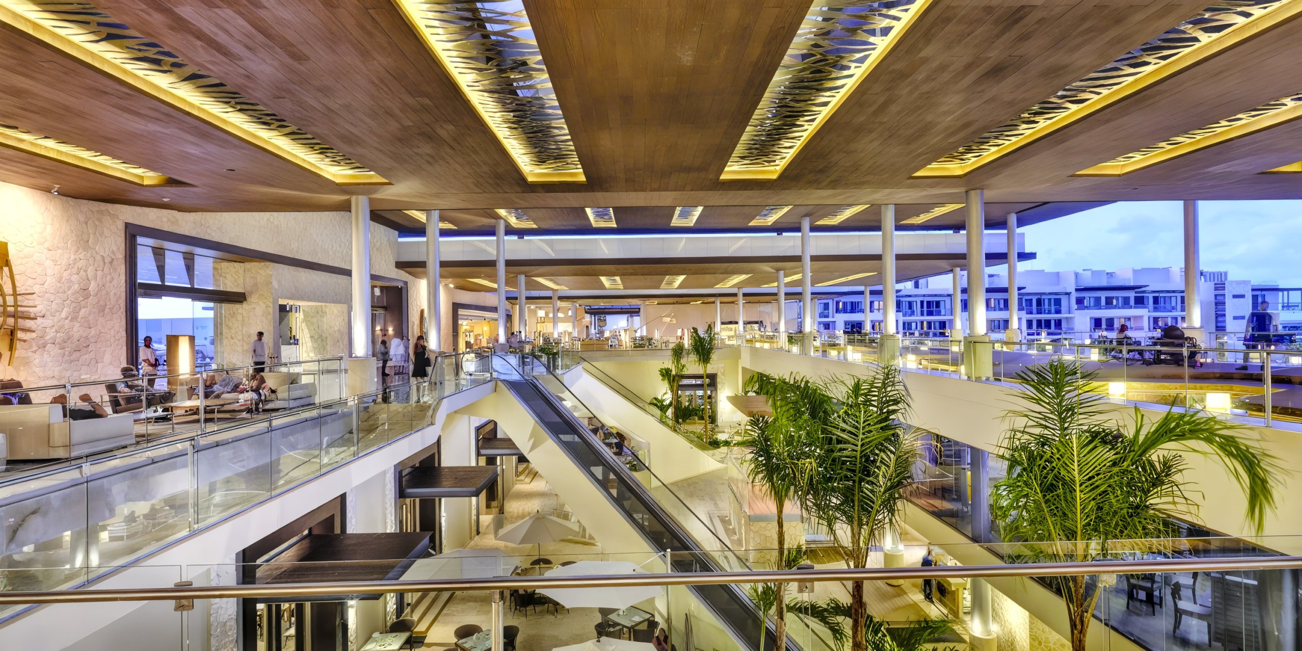 The spacious, modern shopping mall interior, much like the Royalton Riviera Cancun, boasts glass railings, large skylights, and tropical plants. Multiple levels are lively with people walking and shopping.