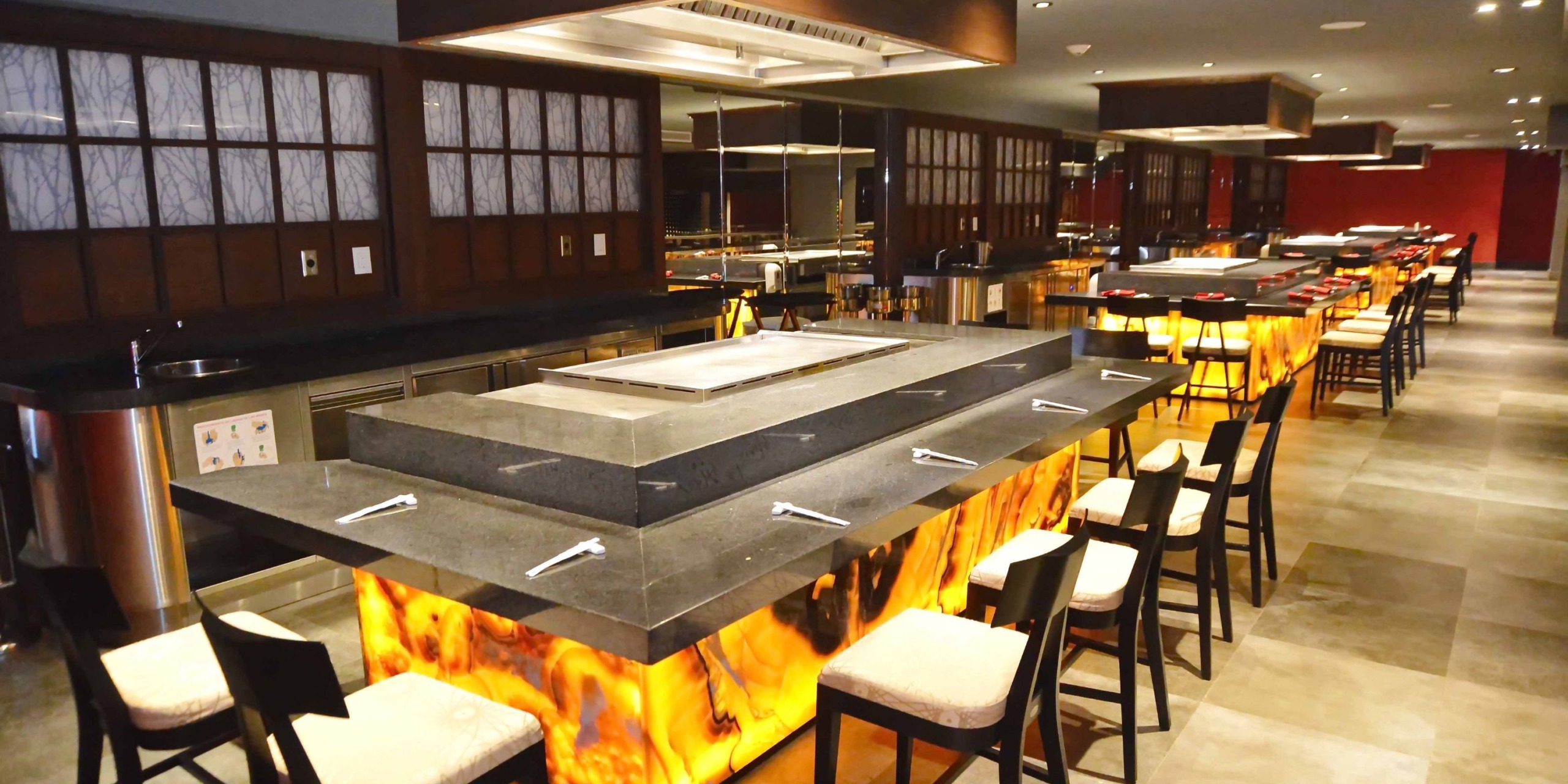 The chic interior of Royalton Riviera Cancun features square tables equipped with built-in grills, complemented by high chairs. Each table is enhanced by an illuminated stone base, while the dark wooden accents add elegance, making it an ideal setting for a fashionable wedding celebration.