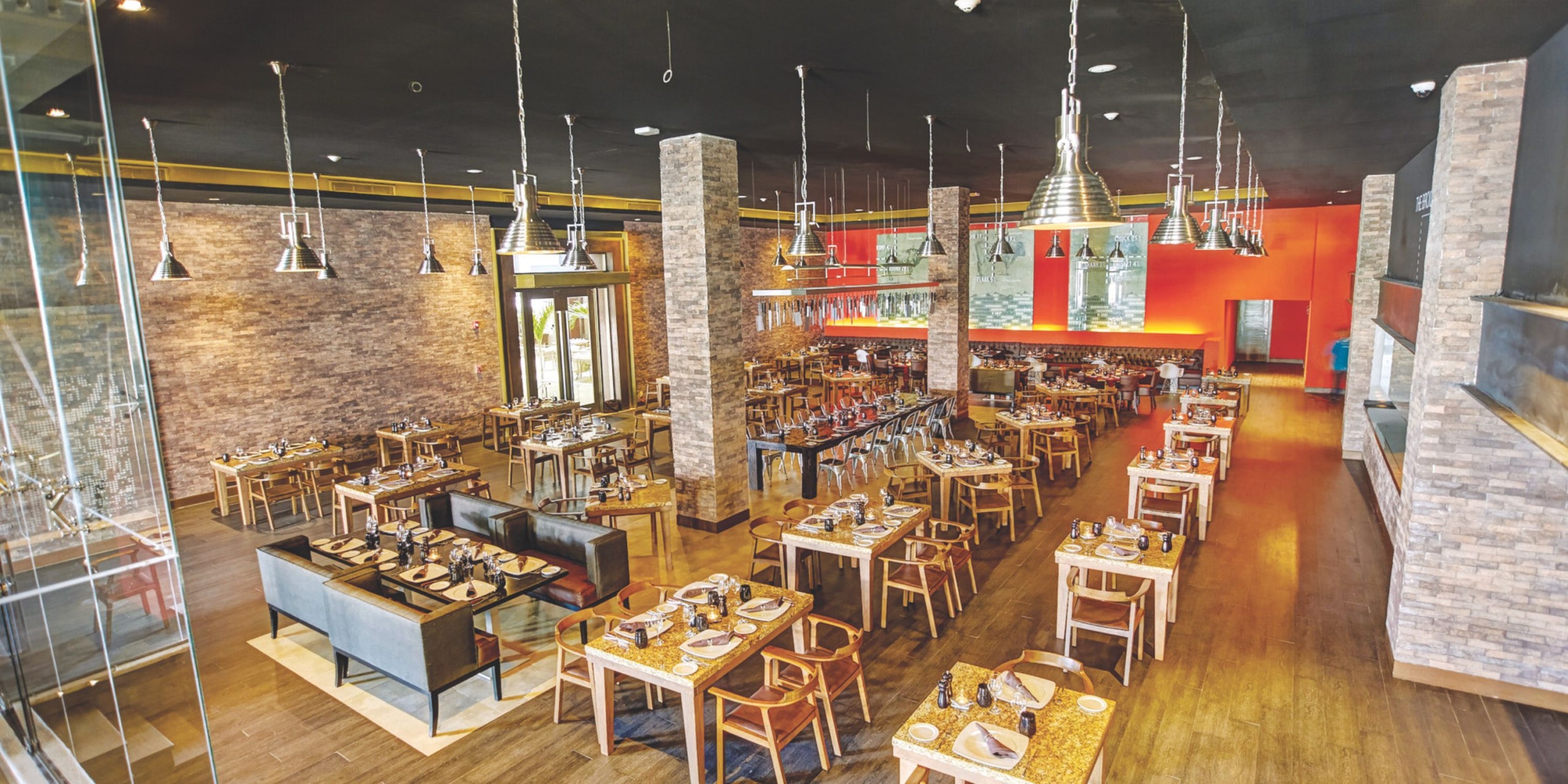 A contemporary restaurant setting with wooden tables and chairs captures the elegance of a Royalton Riviera Cancun wedding. The stone walls and pendant lights enhance the cozy ambiance, while the carefully set tables with plates and cutlery add a sophisticated touch to your dining experience.
