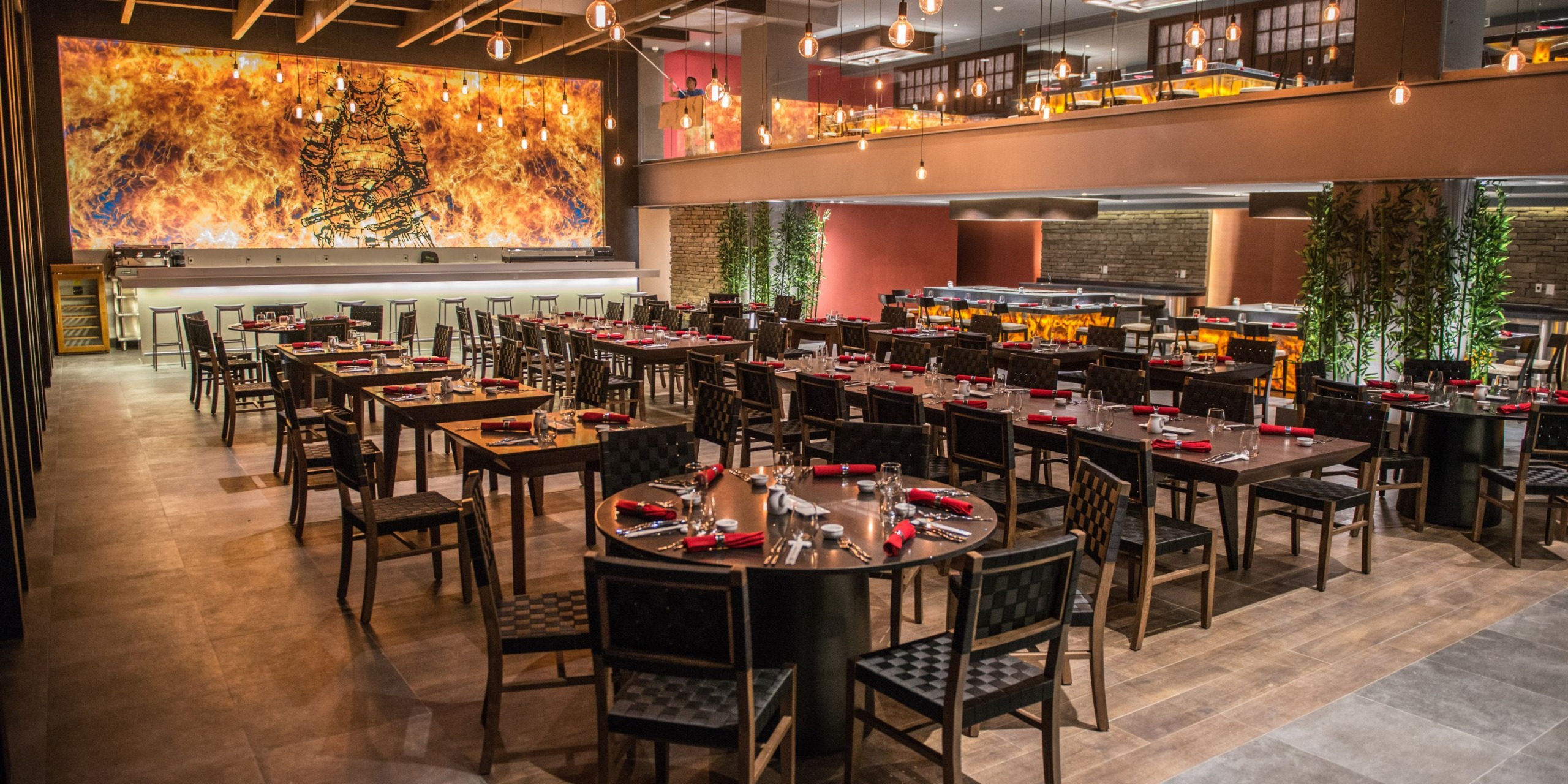 The sleek interior of Royalton Riviera Cancun includes numerous wooden tables and chairs arranged for dining, enhanced by warm lighting and a large, vibrant wall mural that contribute to the ambiance, creating a perfect setting for an unforgettable wedding celebration.