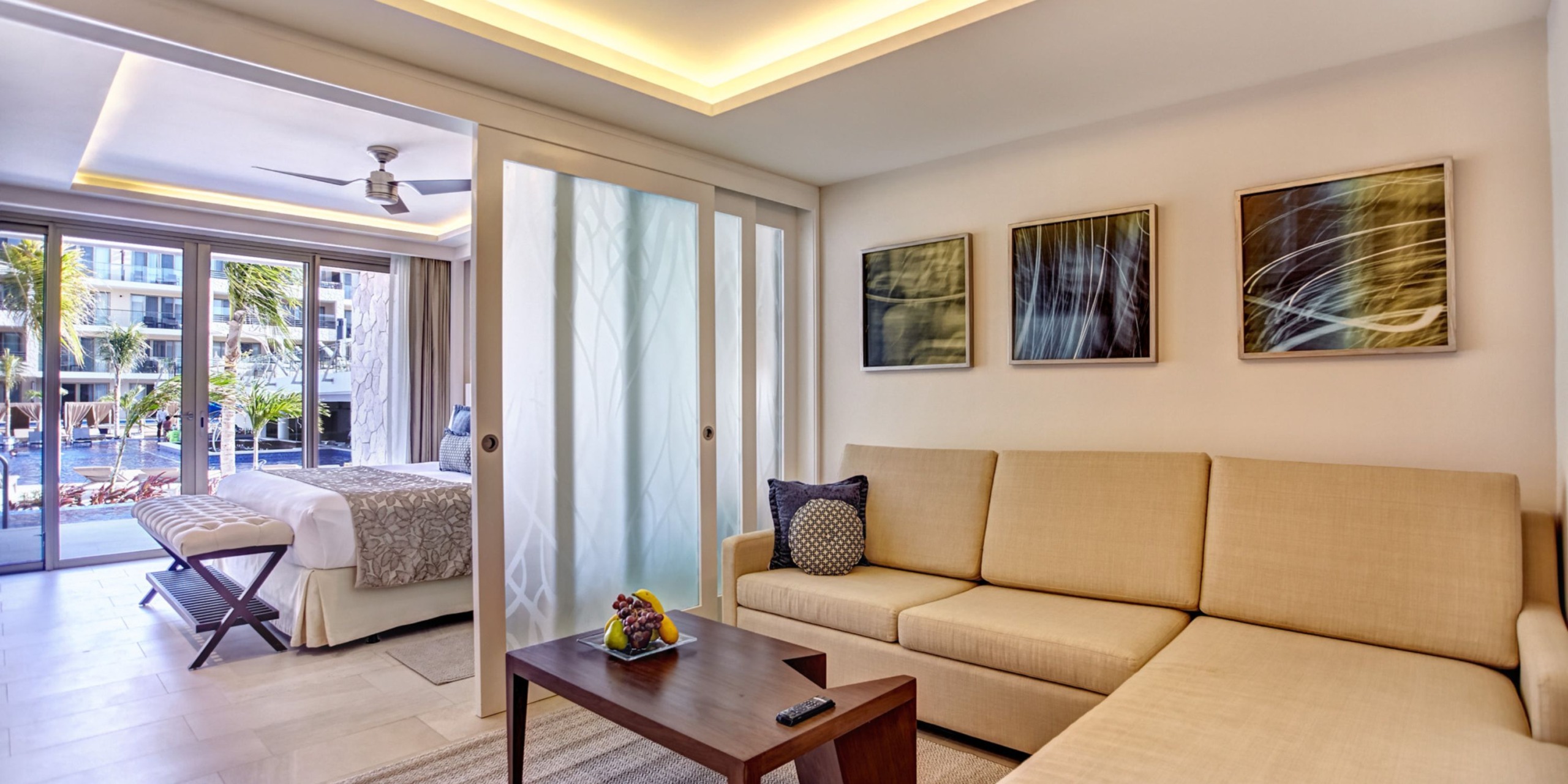 The contemporary hotel room, evocative of a suite at the Royalton Riviera Cancun, includes a beige sectional sofa and a wooden coffee table. A distinct bedroom contains a spacious bed, with sliding glass doors offering a view of the outdoor pool area.