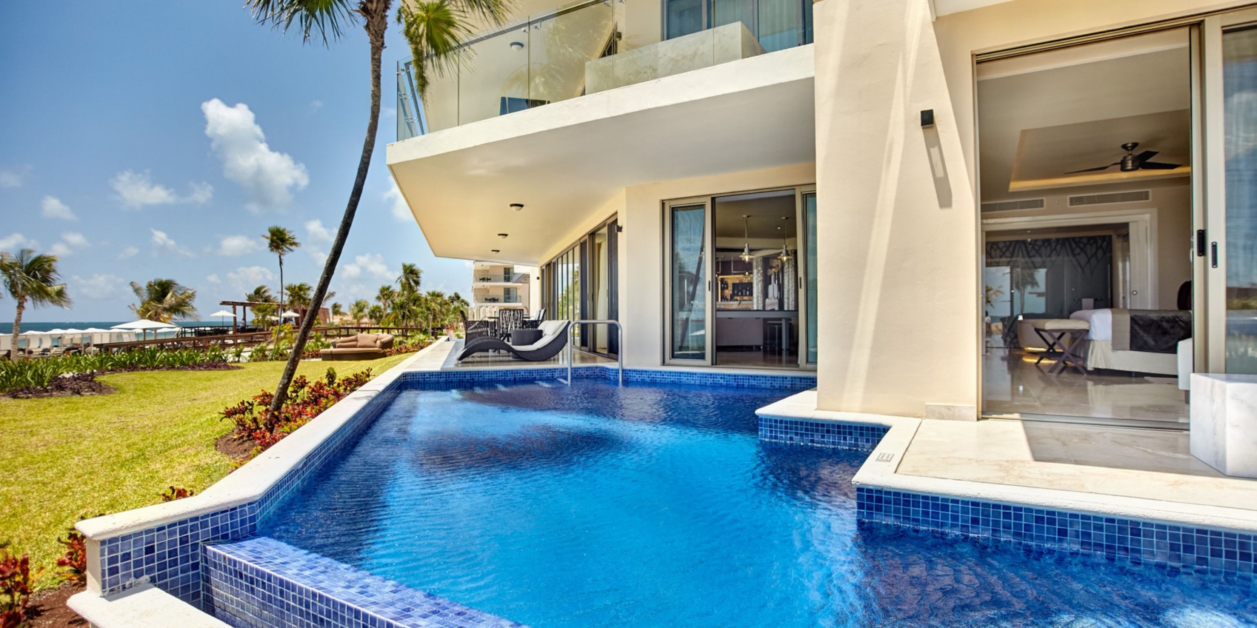 A perfect setting for a wedding at the Royalton Riviera Cancun, featuring a modern building with expansive windows, stylish patio furniture, and lush tropical landscaping all set under a clear blue sky.