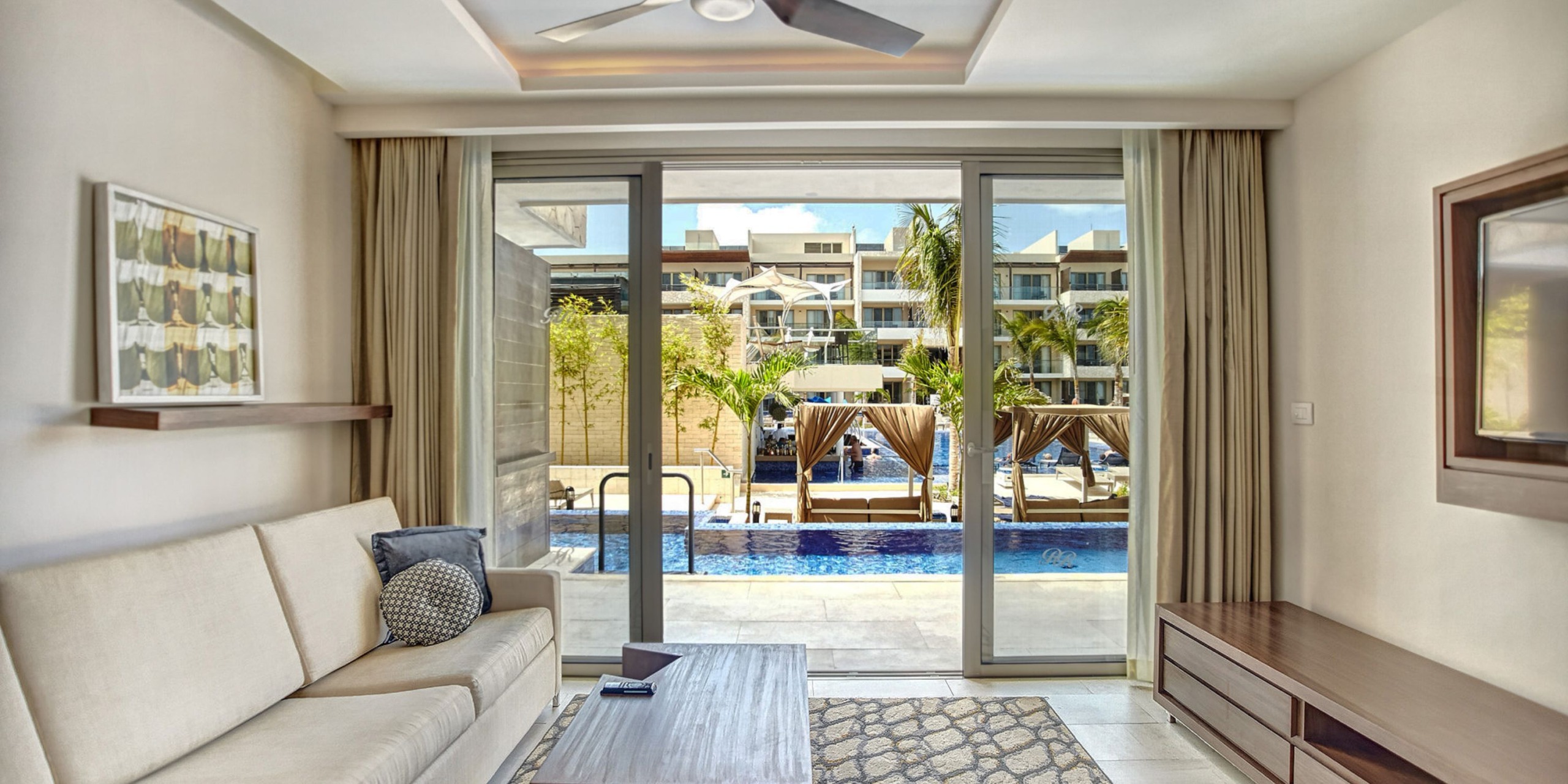 A contemporary living room showcasing a beige sofa, coffee table, and ceiling fan seamlessly opens through glass doors to a poolside view reminiscent of the Royalton Riviera Cancun, complete with cabanas and palm trees gently swaying in the warm breeze.
