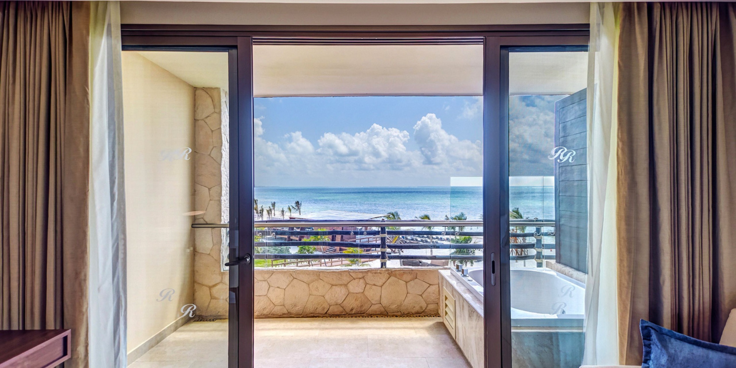 From the balcony of your suite at the Royalton Riviera Cancun, admire the stunning view of the vibrant blue ocean and sky. To your right, a luxurious hot tub beckons you to relax and soak in the tranquil beauty that envelops you.