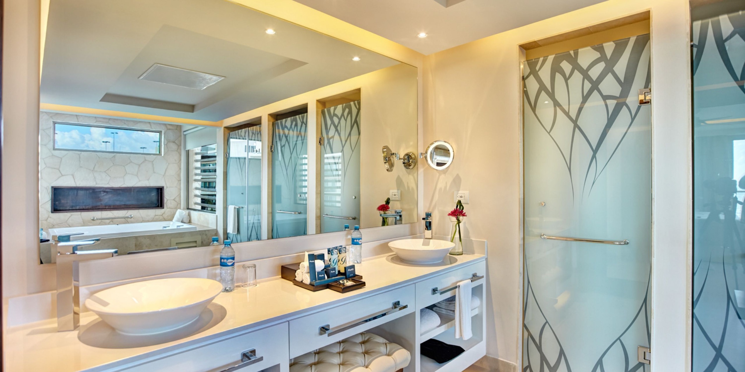 Immerse yourself in a luxury bathroom reminiscent of the Royalton Riviera Cancun, complete with dual sinks on a sleek countertop, an expansive mirror, a frosted glass shower, and a bathtub set against elegantly decorative stone walls.