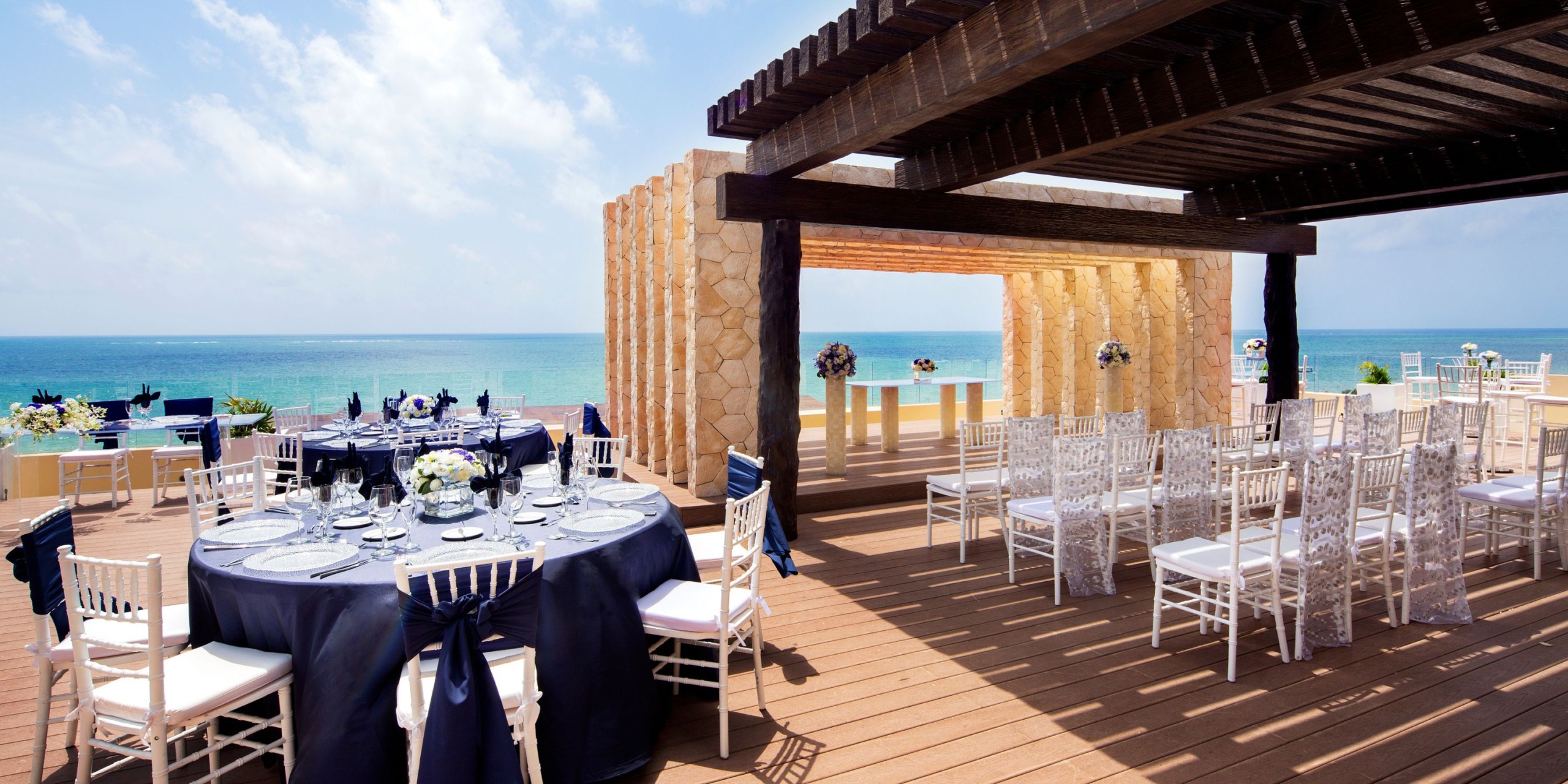 Celebrate your wedding at the Royalton Riviera Cancun with an enchanting outdoor seaside setting. Picture round tables adorned with sophisticated navy tablecloths, elegant white chairs, and a delightful wooden pergola set against the stunning backdrop of the ocean and blue sky.