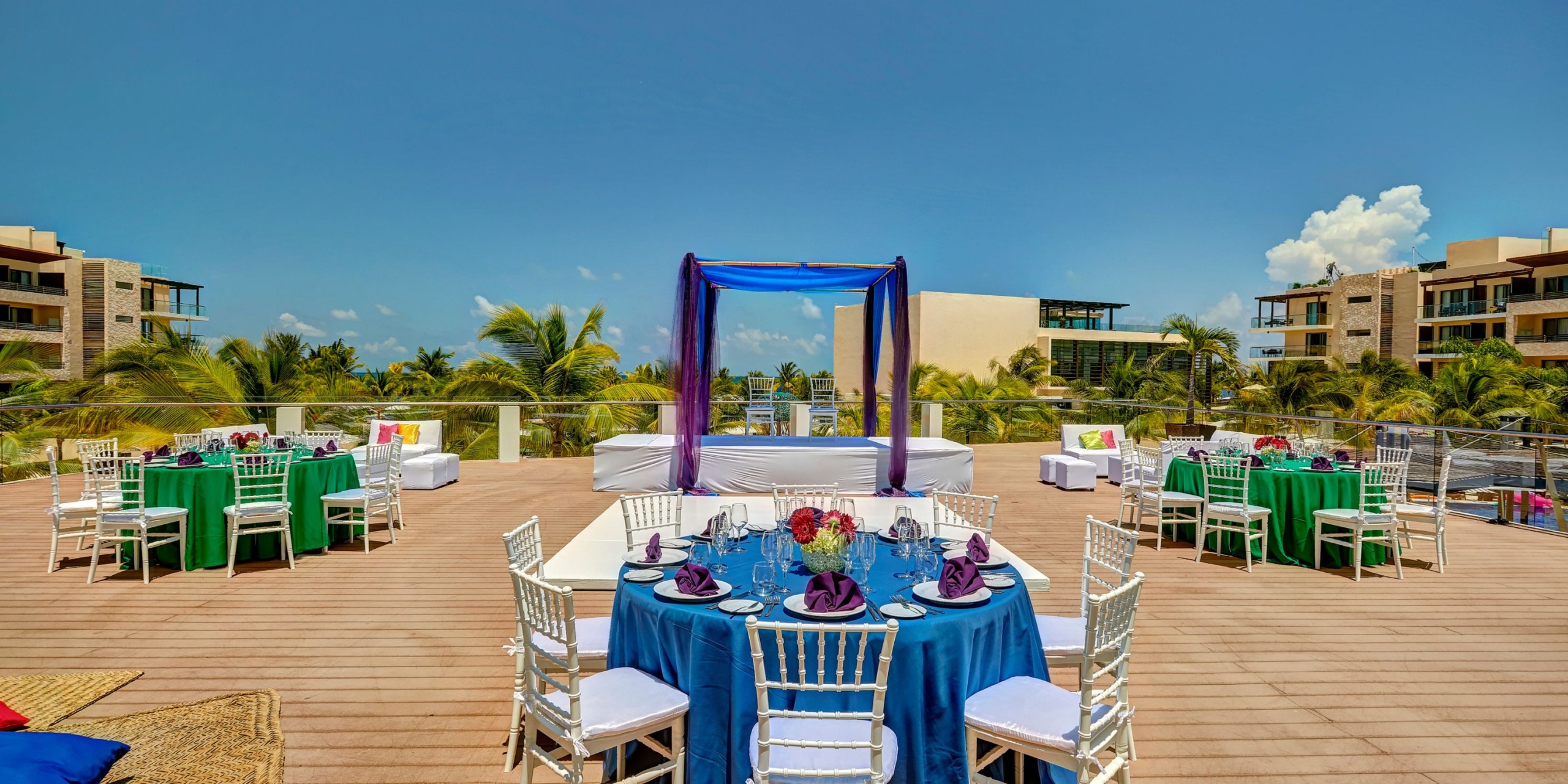 An outdoor patio arrangement for a wedding at the Royalton Riviera Cancun, showcasing round tables adorned with green and blue linens, complemented by white chairs and topped with a canopy against a clear sky backdrop.