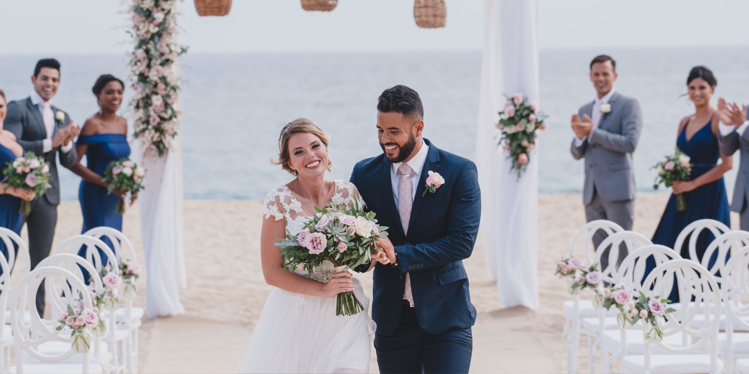 A newlywed couple walks down the aisle on a beach at Sandos Finisterra Los Cabos, surrounded by smiling guests and floral decorations.