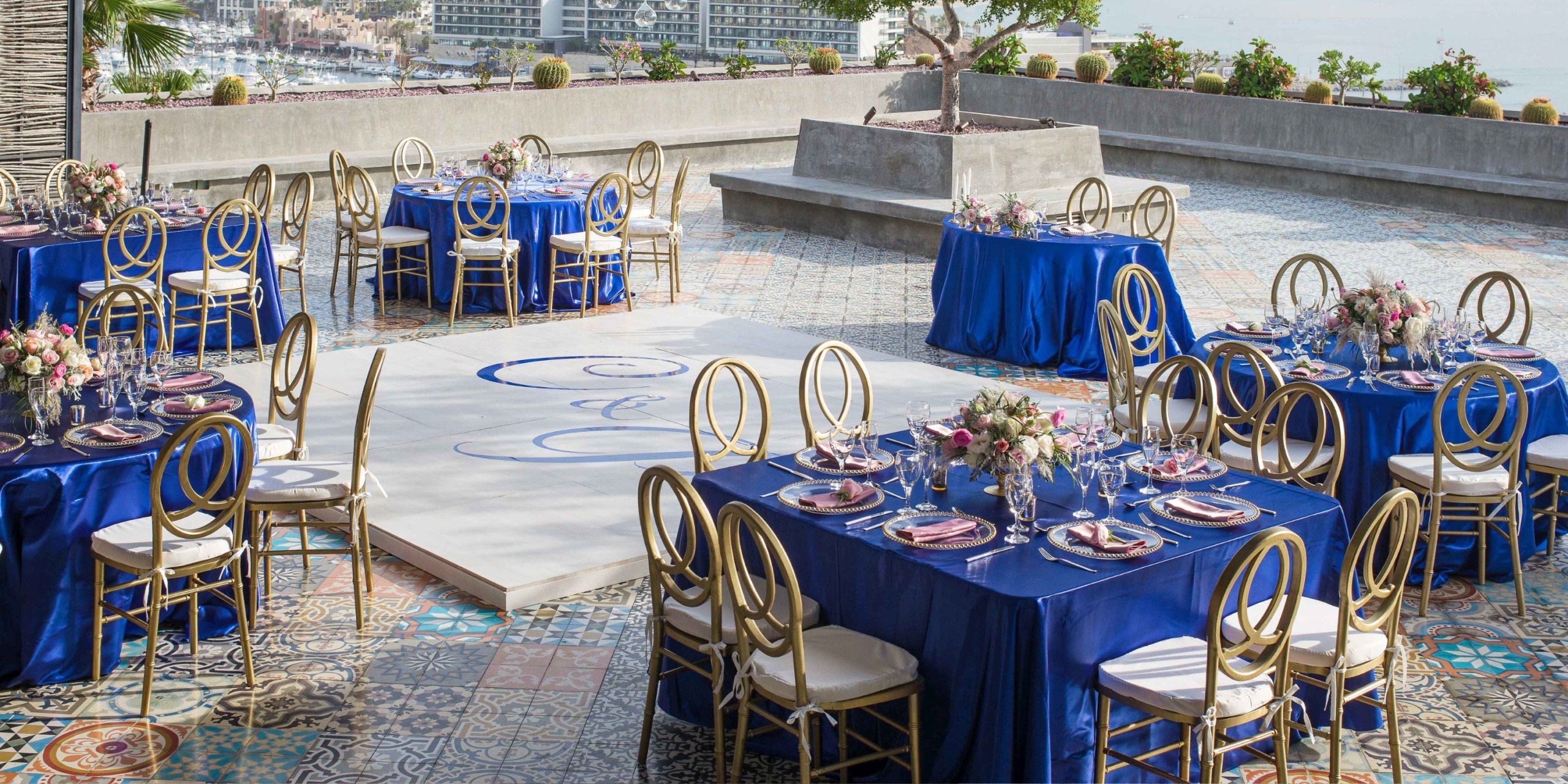 At Sandos Finisterra Los Cabos, the outdoor event features an arrangement with blue tablecloths and gold chairs encircling tables adorned with floral centerpieces. A decorative white dance floor sits prominently in the center, bordered by stylish patterned tiles.