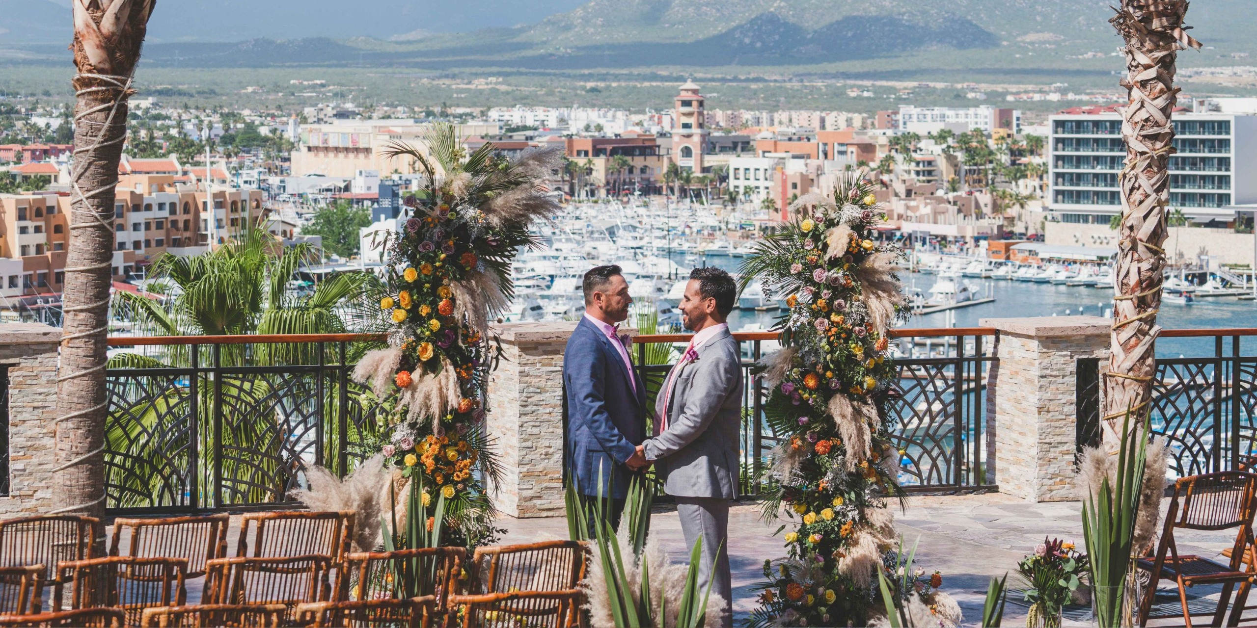 At Sandos Finisterra Los Cabos, two men in suits stand facing each other at an outdoor ceremony adorned with floral arrangements, set against the backdrop of a marina and mountains.