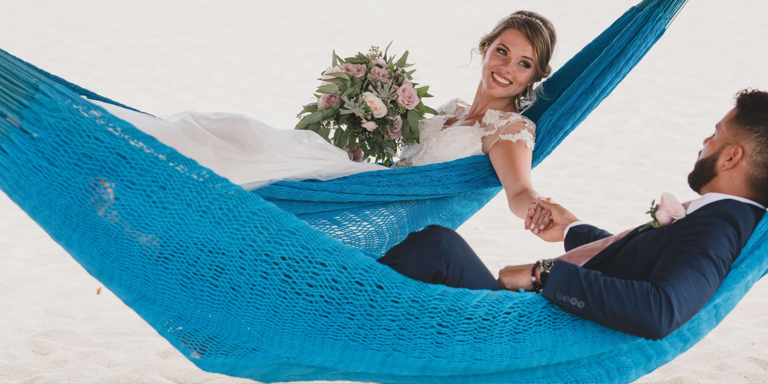 A bride with a bouquet and a groom in a suit relax together on a blue hammock at Sandos Finisterra Los Cabos, holding hands and smiling.