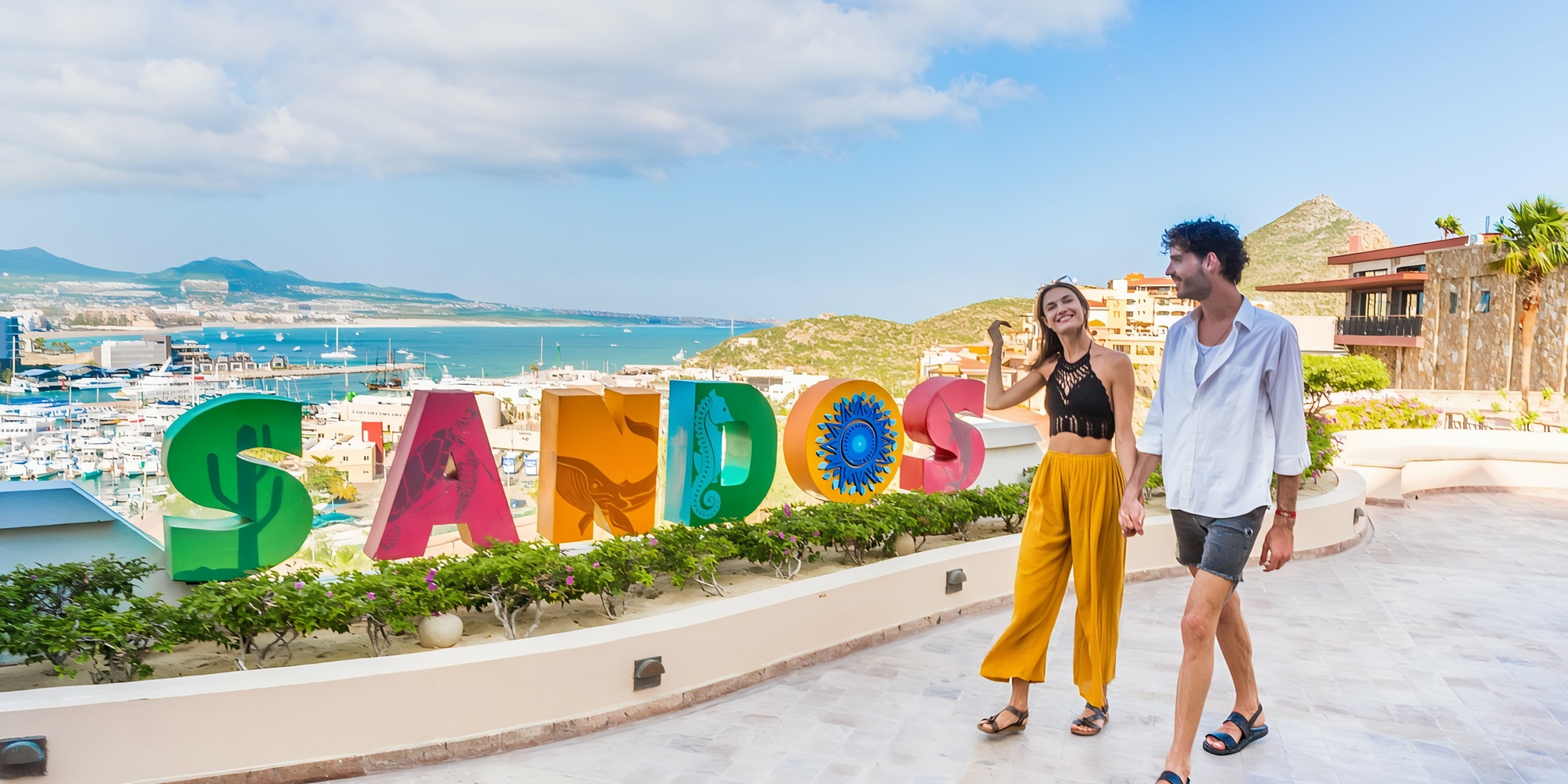 A couple walking and smiling near a colorful Sandos Finisterra Los Cabos sign with a scenic coastal backdrop.
