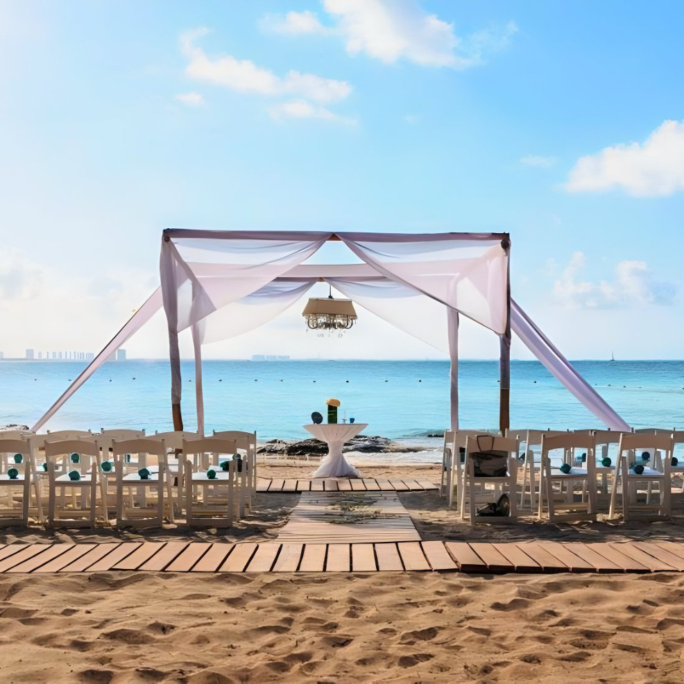 A destination wedding setup on a beach with an elegant white canopy, white chairs arranged on the sand, and a scenic ocean backdrop under a clear blue sky.