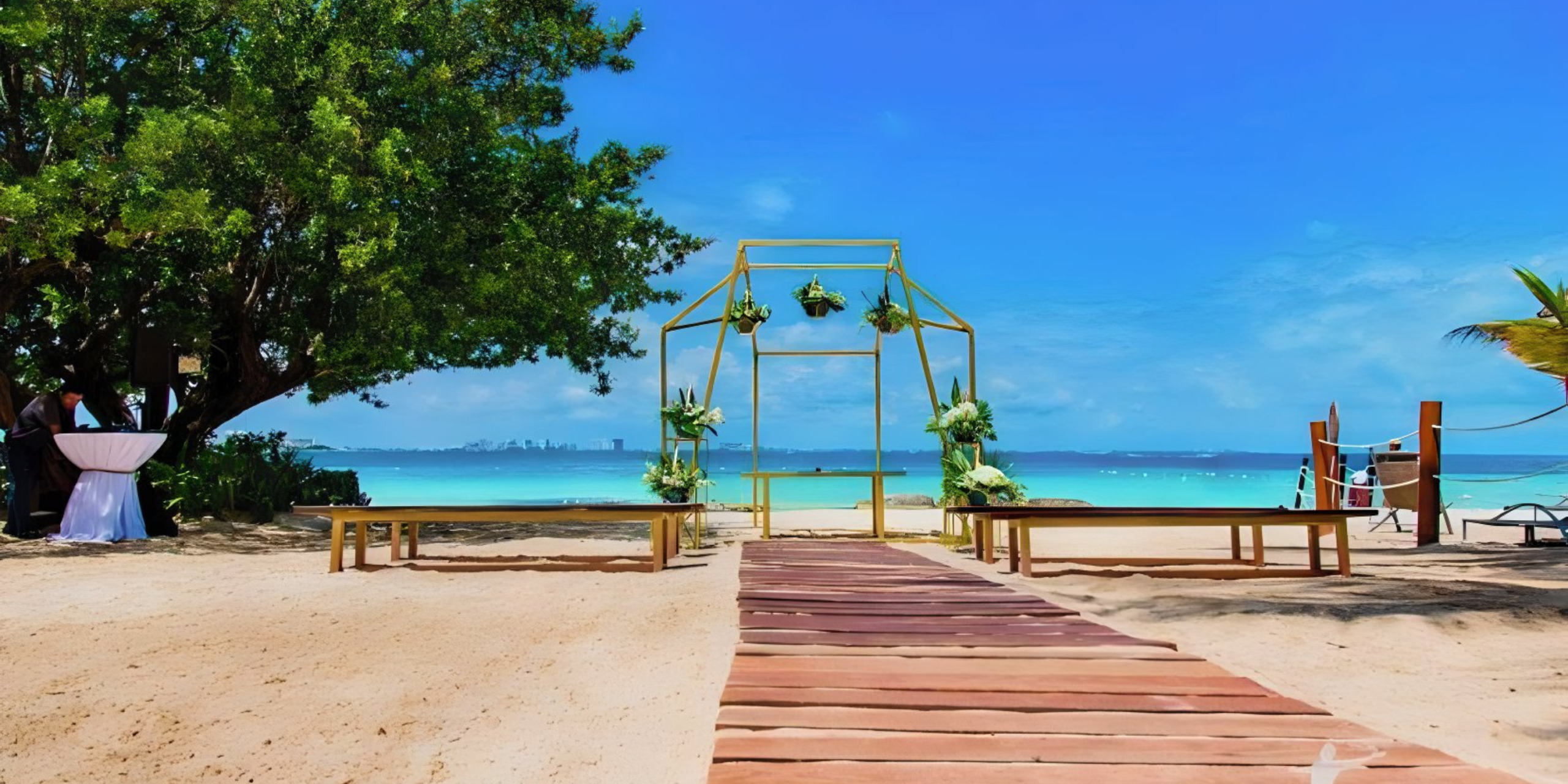 A destination beach wedding setup with a wooden walkway leading to a floral-decorated arch, benches on either side, and a clear blue sky backdrop.