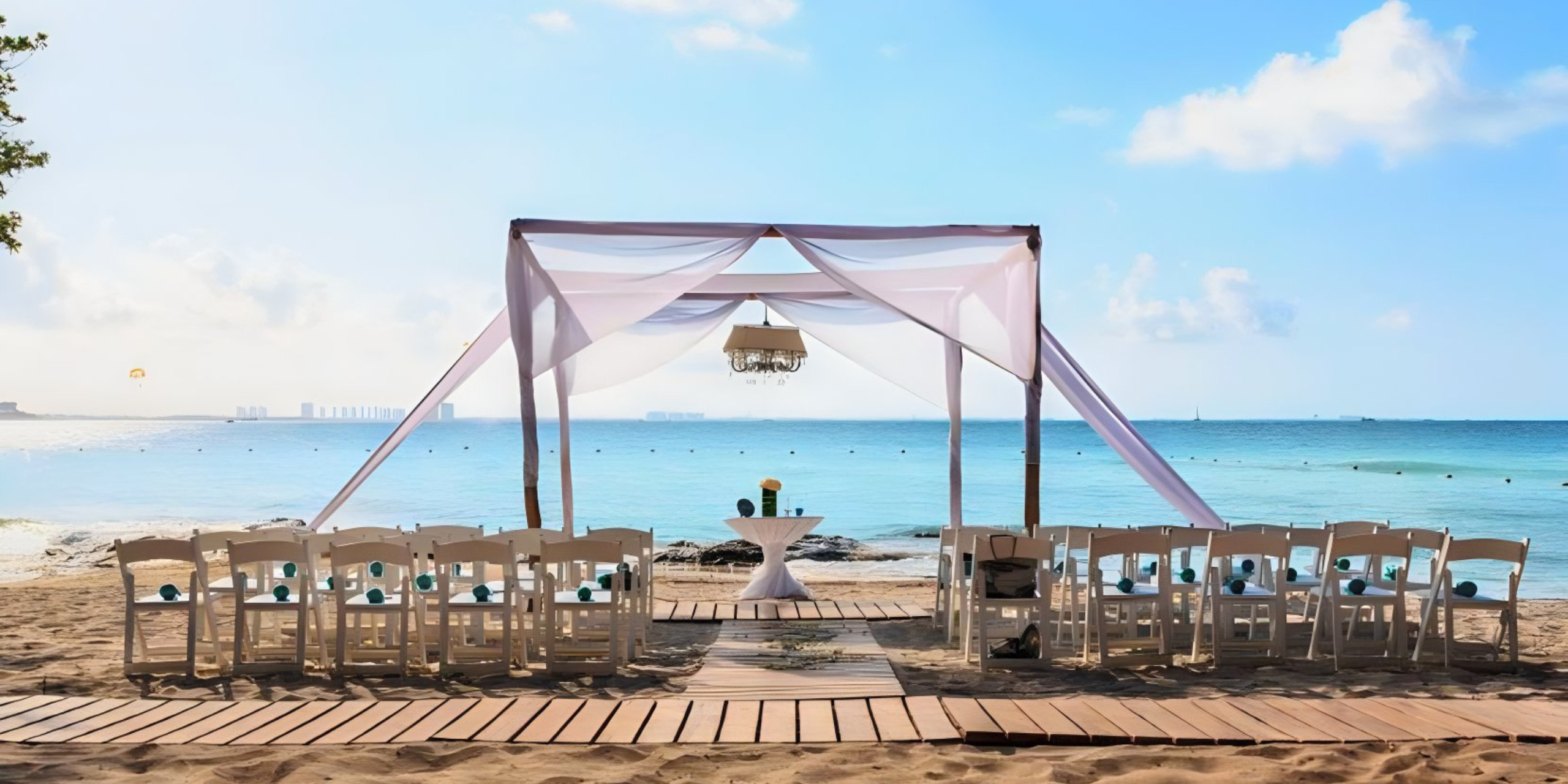 A destination wedding setup on the beach with a white canopy, chairs, and a wooden aisle leading to the ocean under a clear blue sky.