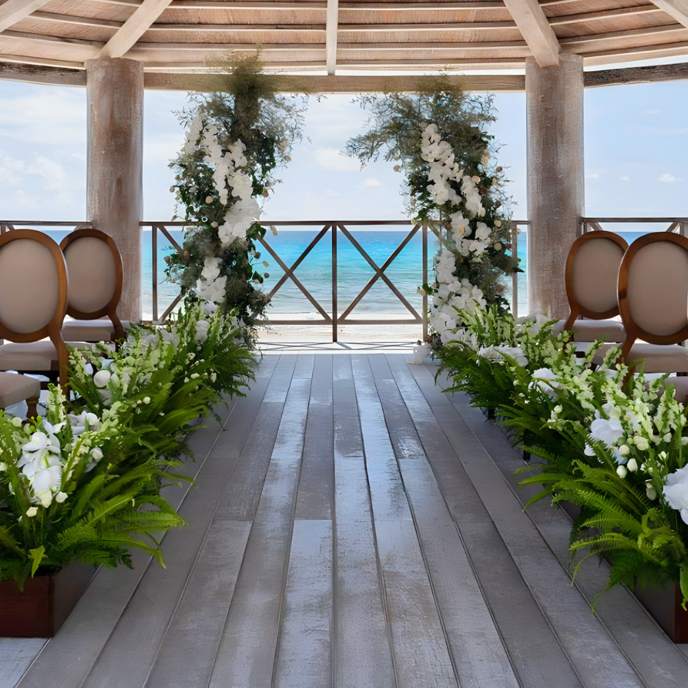 A destination wedding venue decorated with white flowers and greenery under a wooden pergola, overlooking the ocean.