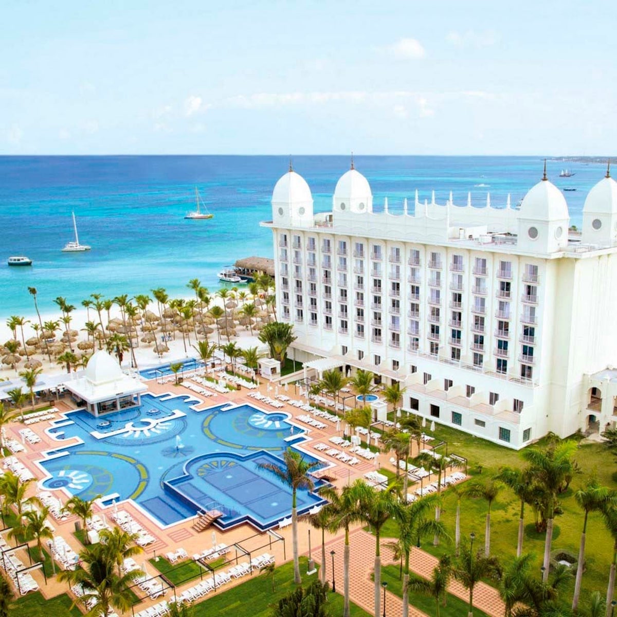 A luxurious beachfront Hotel Riu Palace Antillas, perfect for a destination wedding, with white domes, a large swimming pool, and several boats on the clear blue ocean in the background.