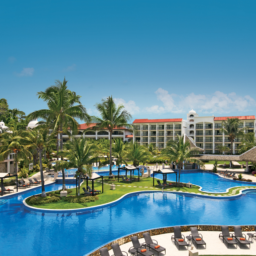 A tropical resort, Dreams Playa Bonita, a perfect destination wedding venue, with a large blue swimming pool surrounded by lounge chairs, palm trees, and a multi-storey hotel building under a clear blue sky.