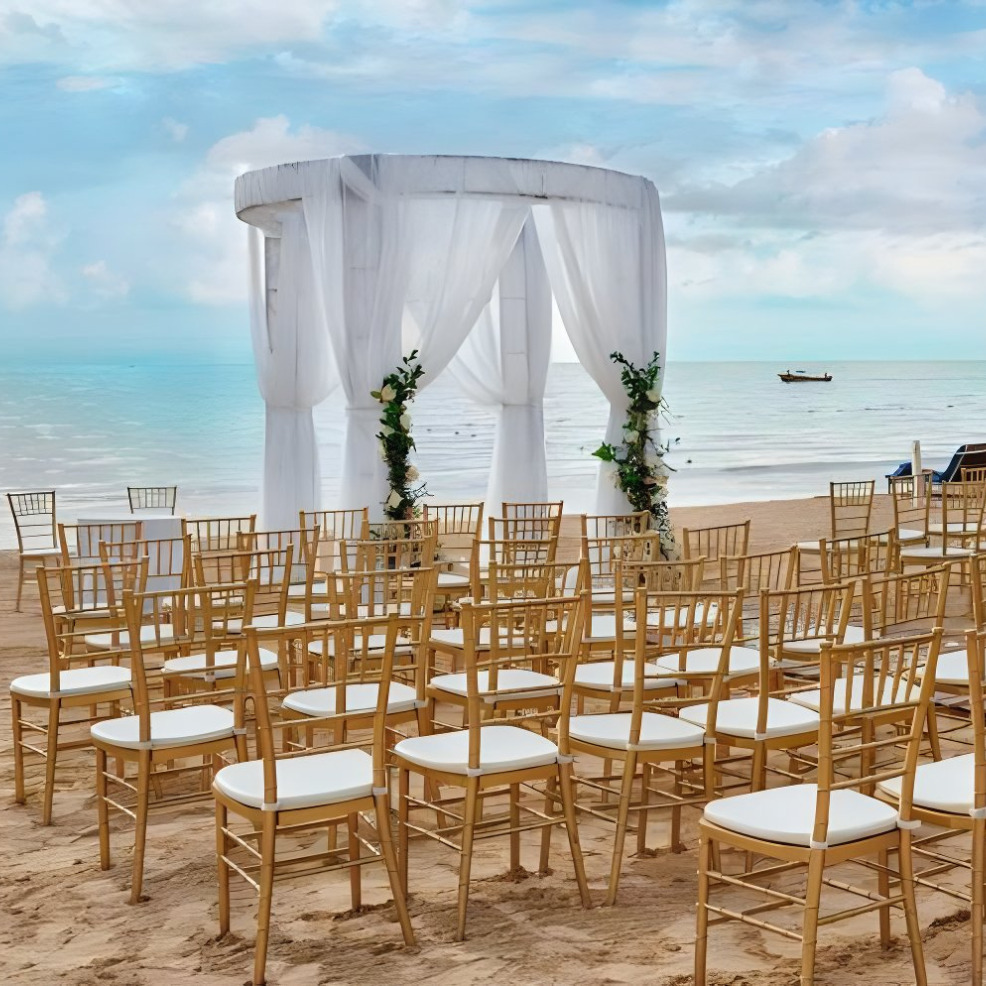 A destination beach wedding setup with a white draped gazebo and rows of wooden chairs on a sandy shore, ocean and a boat in the background.