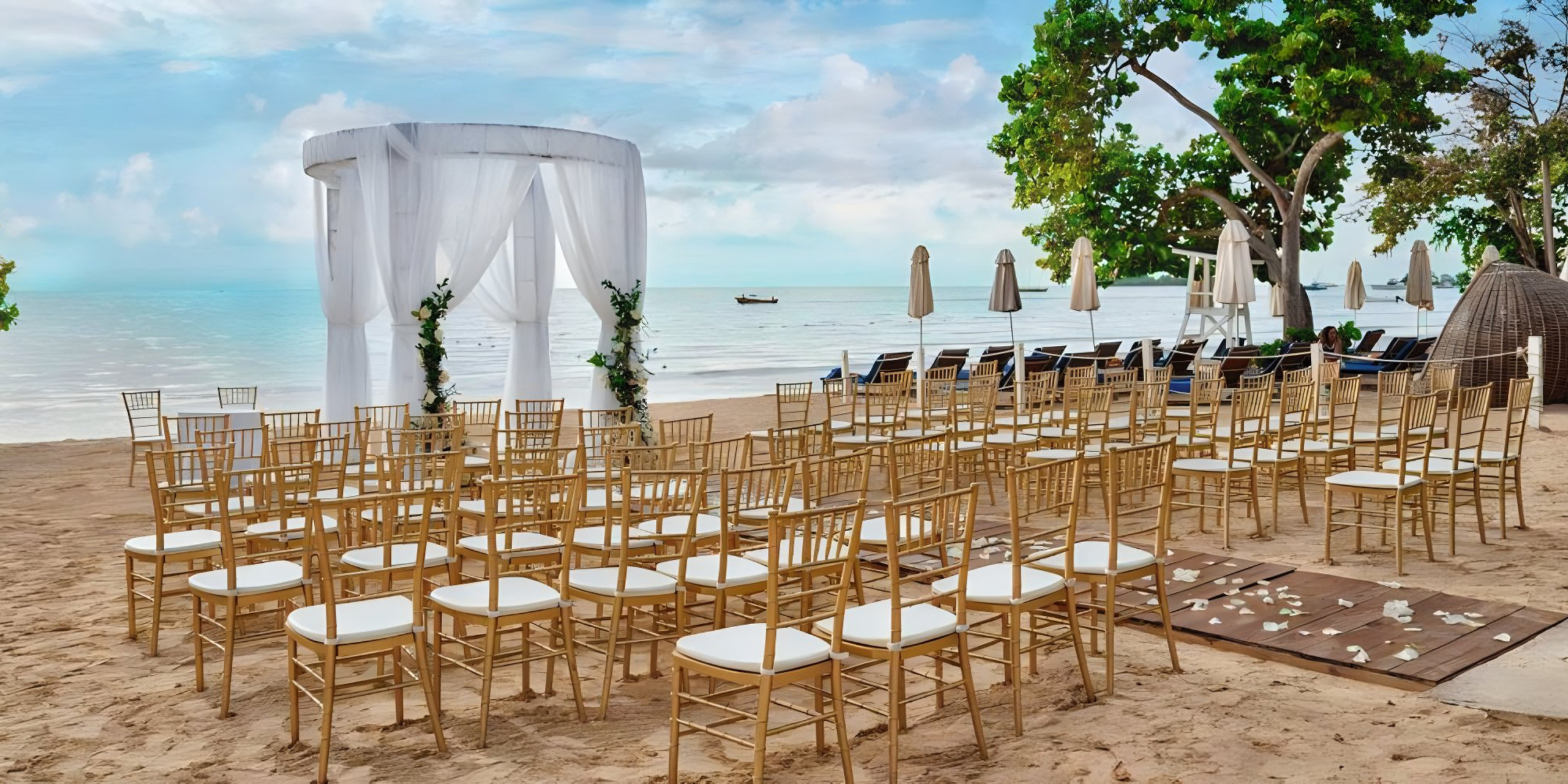 Beach destination wedding setup with white chairs, a floral arch, and a clear view of the ocean under a sunny sky.